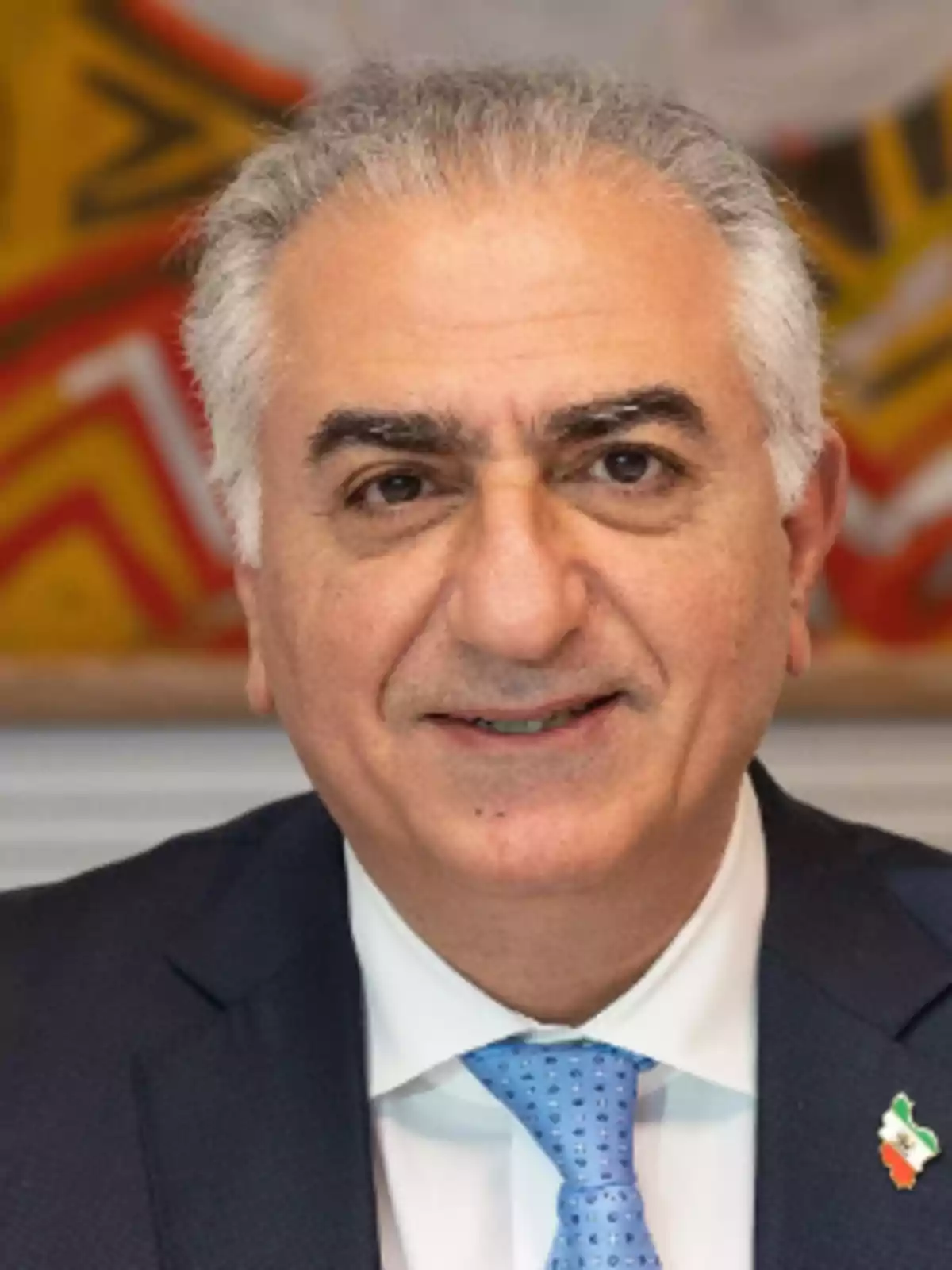 Hombre de cabello canoso con traje azul y corbata celeste sonriendo frente a una pintura colorida Hombre de cabello canoso con traje azul y corbata celeste sonriendo frente a una pintura colorida