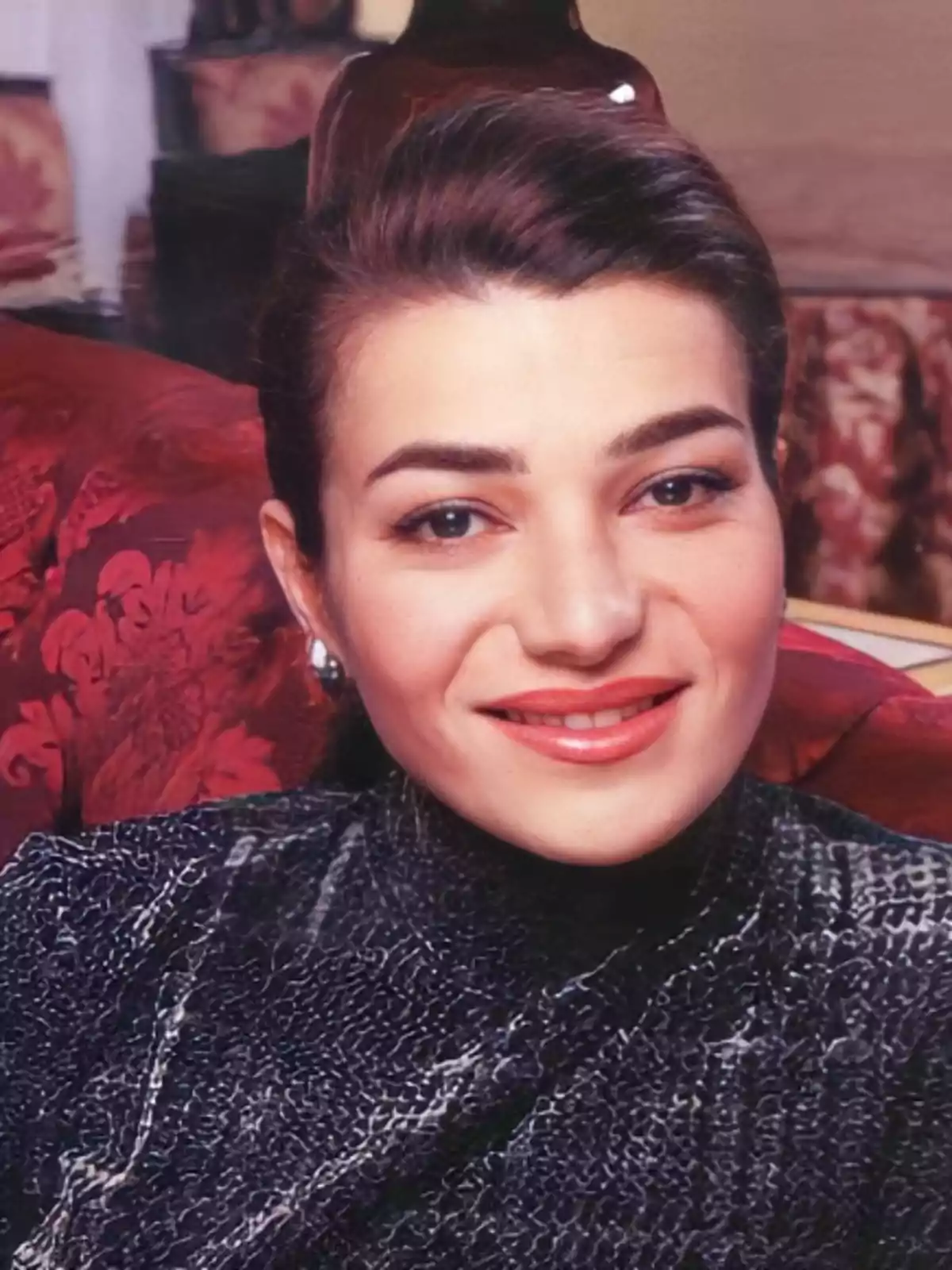Mujer joven de cabello oscuro recogido, con suéter de textura gris y fondo de sillón rojo, sonriendo a la cámara Mujer joven de cabello oscuro recogido, con suéter de textura gris y fondo de sillón rojo, sonriendo a la cámara