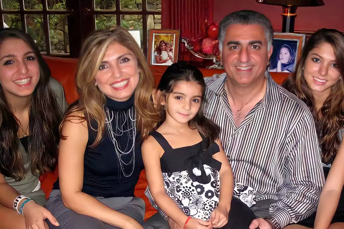 Familia sentada en un sofá sonriendo a la cámara con fotos familiares al fondo Familia sentada en un sofá sonriendo a la cámara con fotos familiares al fondo
