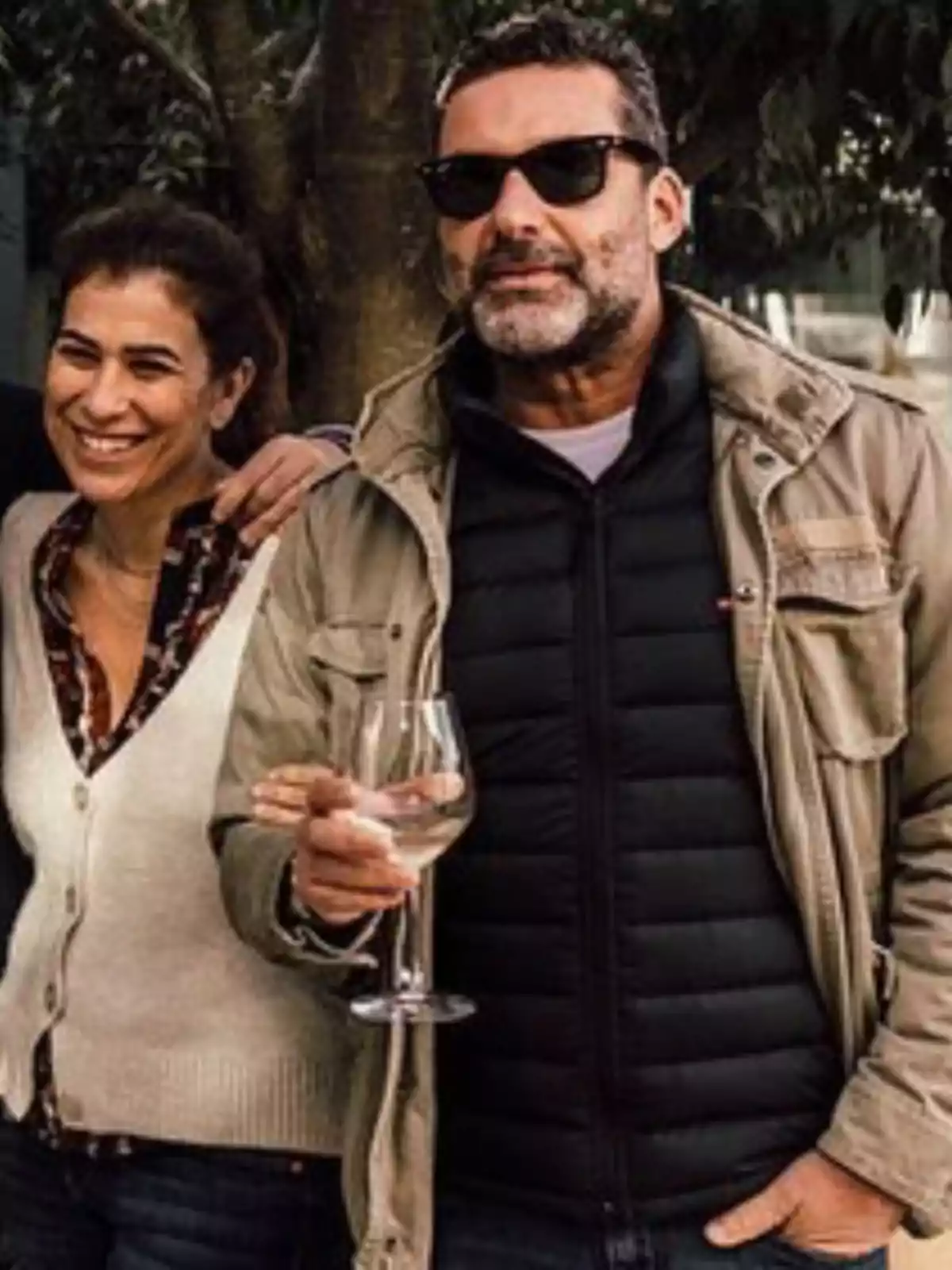 Dos personas sonríen al aire libre, una de ellas lleva gafas de sol y sostiene una copa de vino blanco. Dos personas sonríen al aire libre, una de ellas lleva gafas de sol y sostiene una copa de vino blanco.