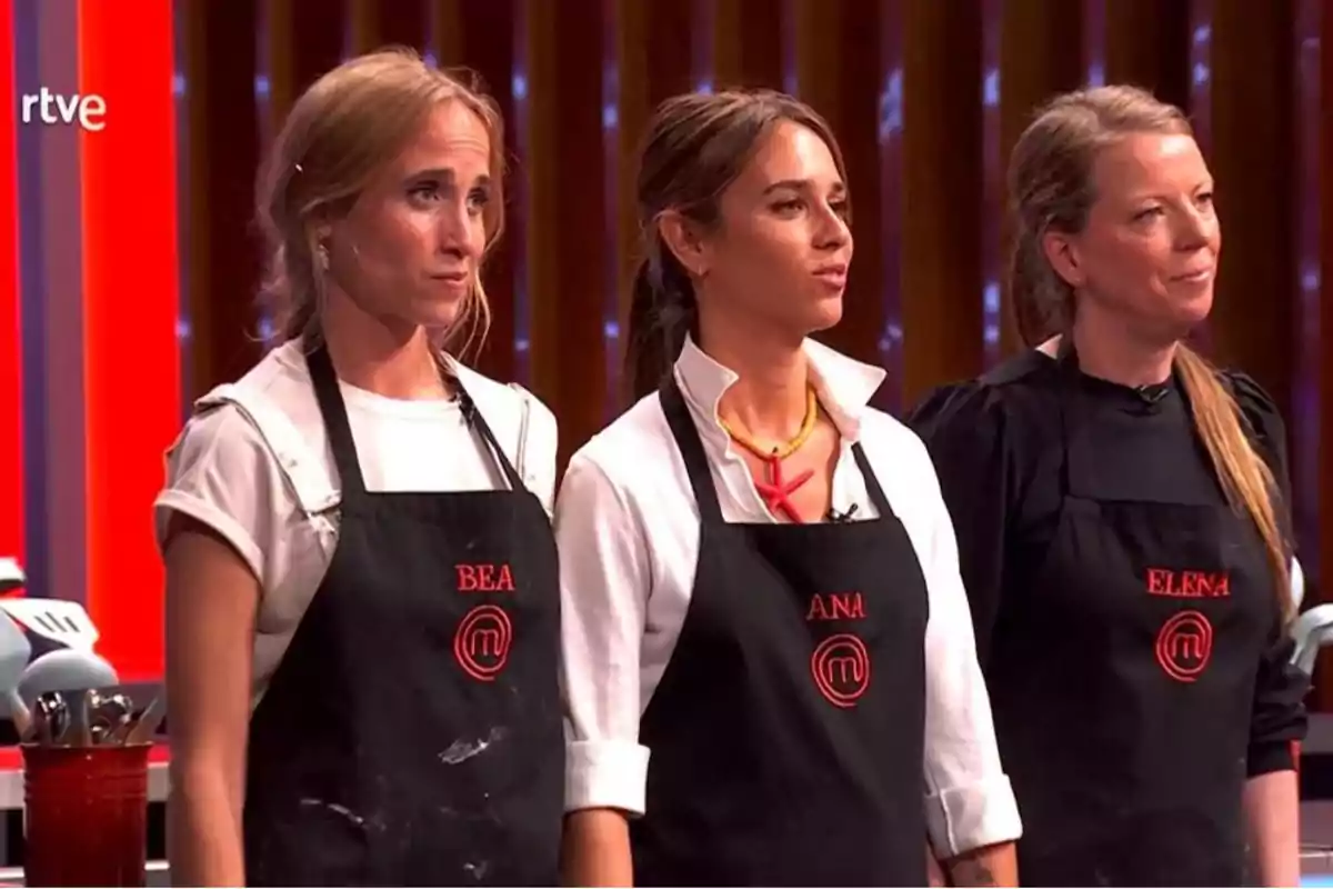 Tres concursantes de un programa de cocina con delantales negros y nombres bordados en rojo están de pie en una cocina iluminada Tres concursantes de un programa de cocina con delantales negros y nombres bordados en rojo están de pie en una cocina iluminada