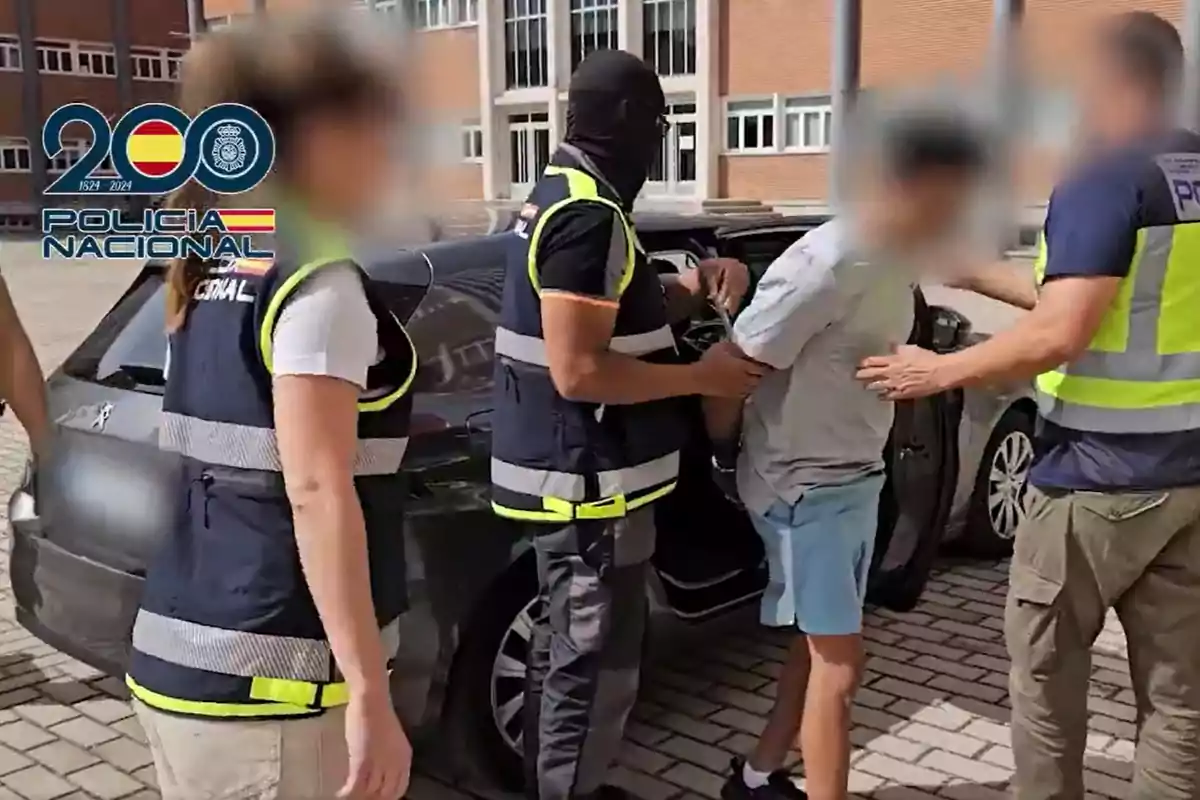 Varios agentes de la Policía Nacional detienen a una persona junto a un coche en la vía pública frente a un edificio, todos llevan chalecos identificativos y algunos rostros están difuminados.