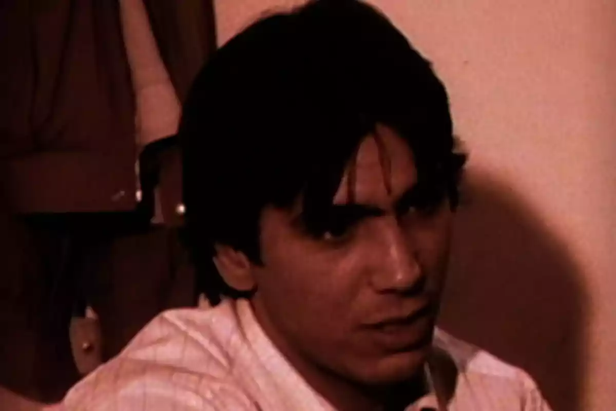 Hombre de cabello oscuro con expresión seria en una habitación con paredes claras y ropa colgada al fondo Hombre de cabello oscuro con expresión seria en una habitación con paredes claras y ropa colgada al fondo