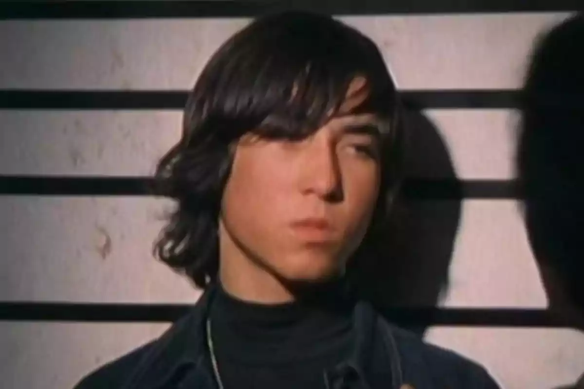 Joven de cabello largo y oscuro frente a una pared con líneas horizontales, mirando hacia un lado con expresión seria Joven de cabello largo y oscuro frente a una pared con líneas horizontales, mirando hacia un lado con expresión seria