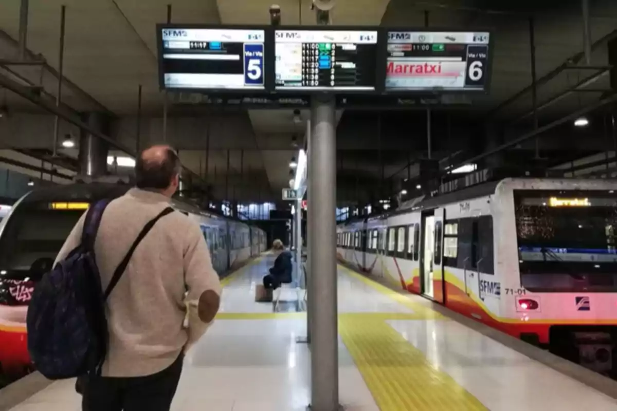 Un hombre de pie en un andén de estación de tren observa los paneles informativos mientras un tren está detenido y otra persona está sentada esperando.