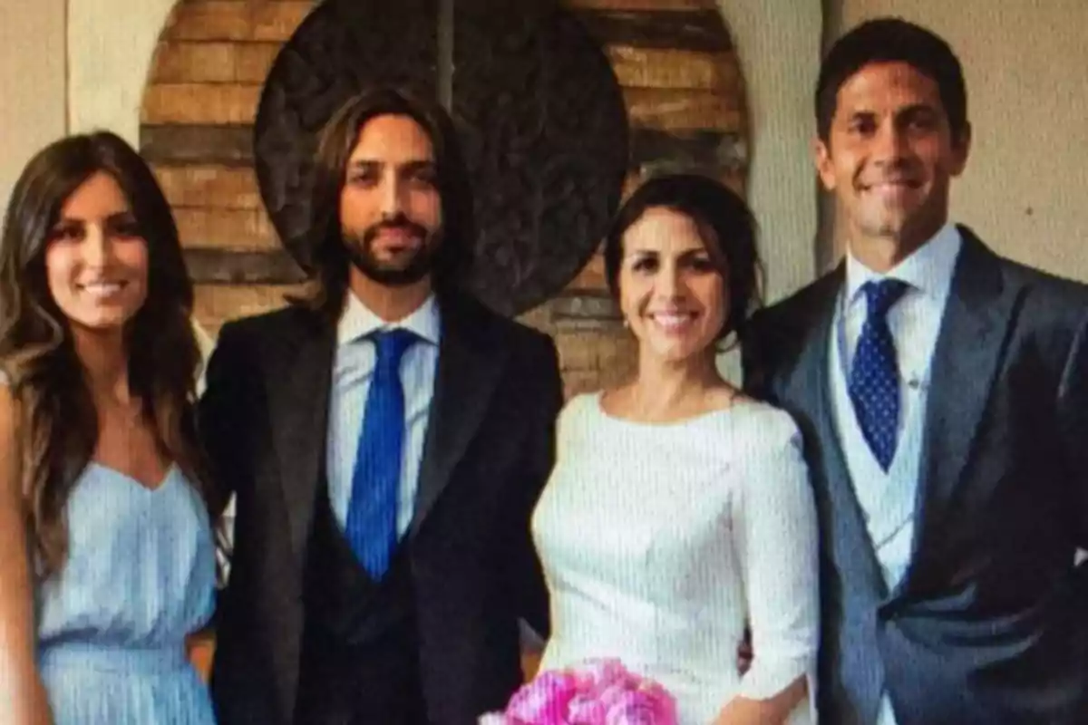 Cuatro personas posan sonrientes en lo que parece ser una boda, dos hombres con traje y dos mujeres, una de ellas con vestido blanco y ramo de flores.