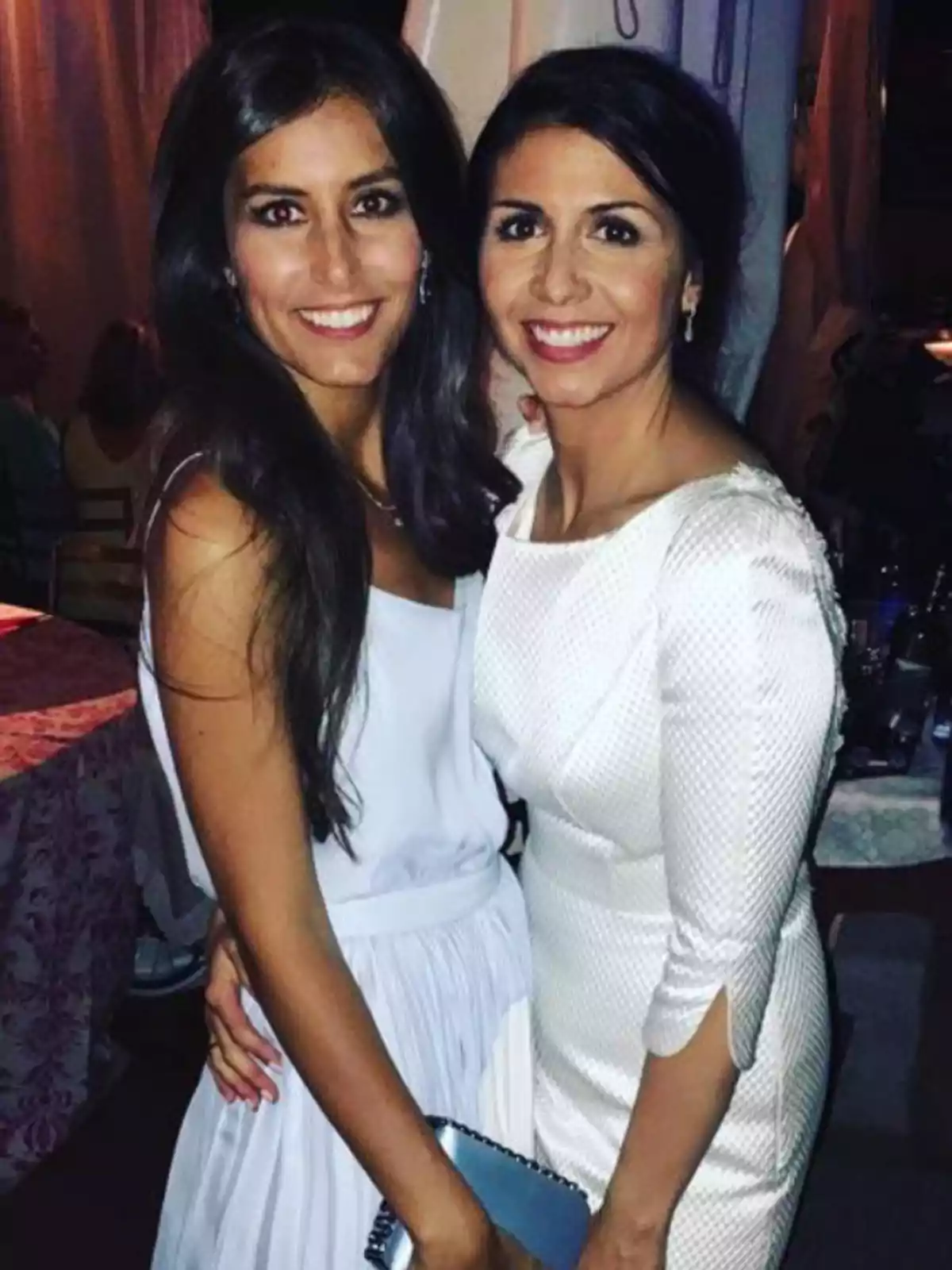 Dos mujeres sonríen abrazadas mientras posan para una foto en un evento elegante, ambas llevan vestidos claros y el ambiente es de fiesta.