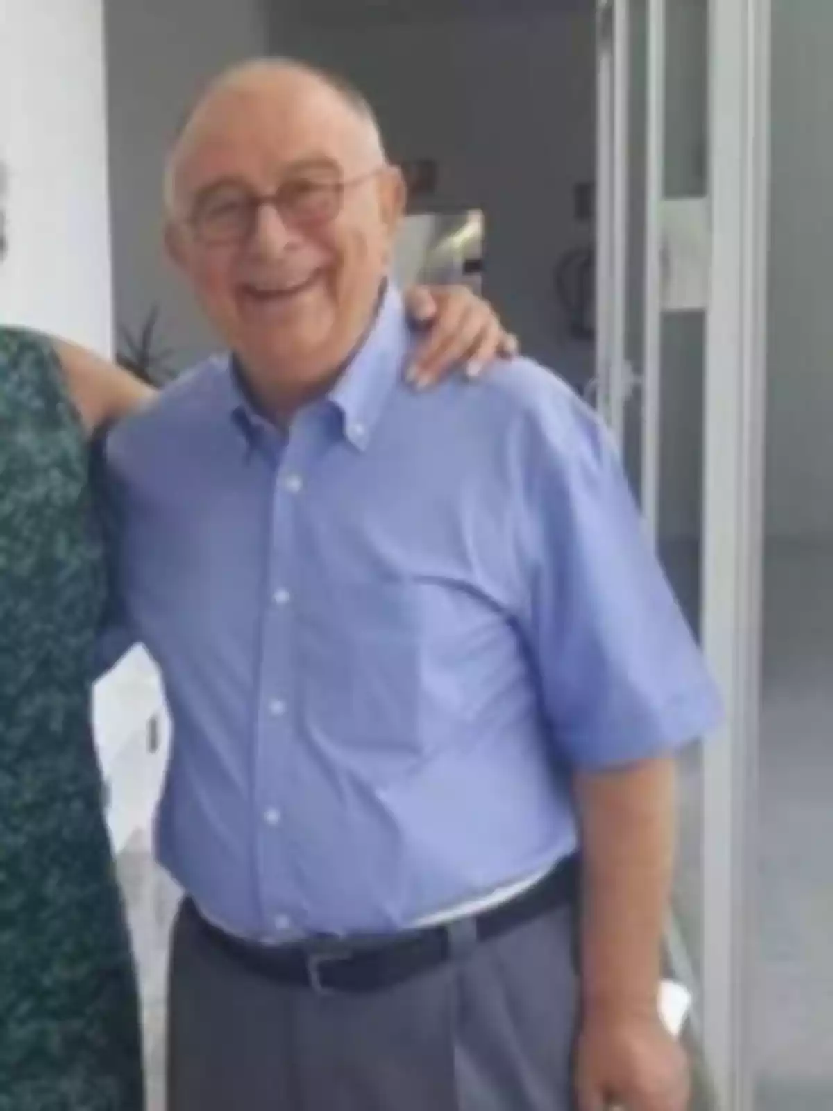 Hombre mayor sonriente con gafas y camisa azul de pie junto a otra persona en un entorno interior luminoso Hombre mayor sonriente con gafas y camisa azul de pie junto a otra persona en un entorno interior luminoso