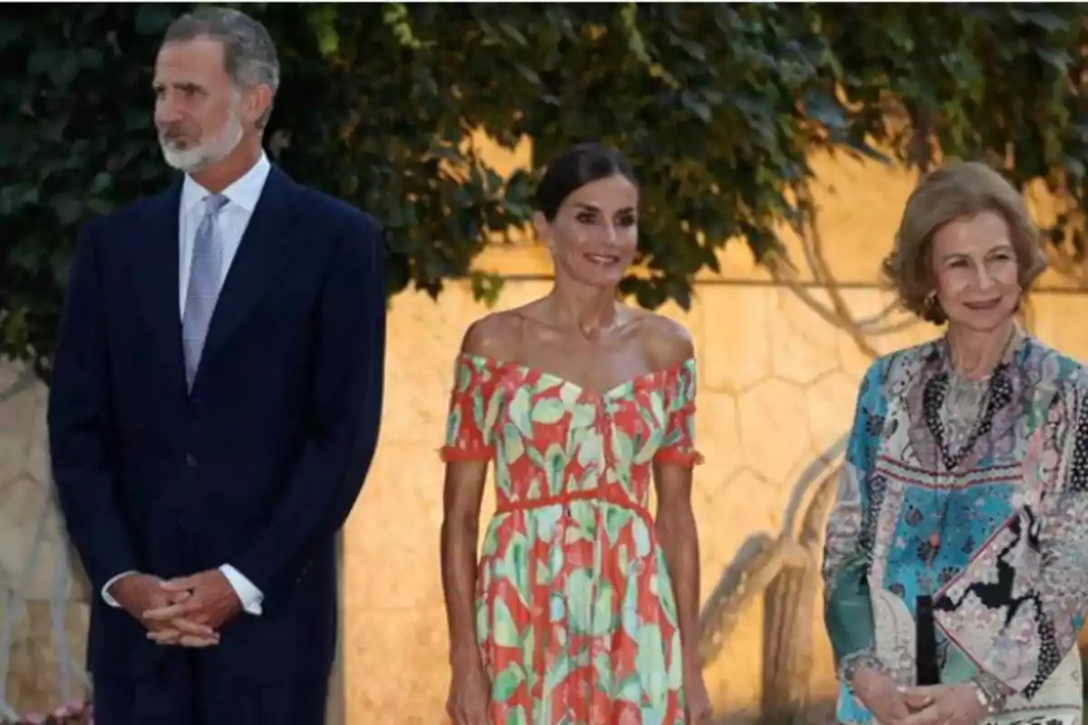 Tres personas elegantemente vestidas posan al aire libre frente a un fondo de vegetación y una pared iluminada Tres personas elegantemente vestidas posan al aire libre frente a un fondo de vegetación y una pared iluminada