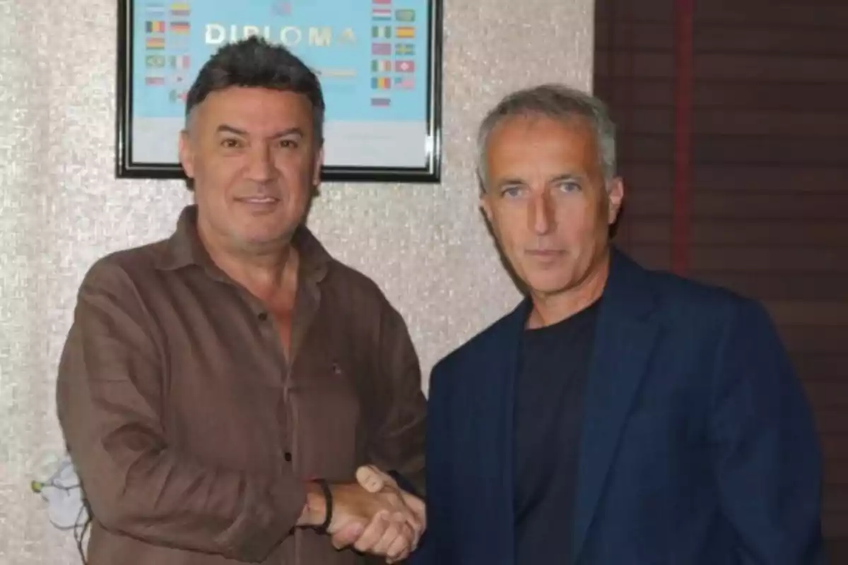 Dos hombres se dan la mano y miran a la cámara, uno lleva camisa marrón y el otro saco azul, al fondo hay un diploma enmarcado en la pared. Dos hombres se dan la mano y miran a la cámara, uno lleva camisa marrón y el otro saco azul, al fondo hay un diploma enmarcado en la pared.