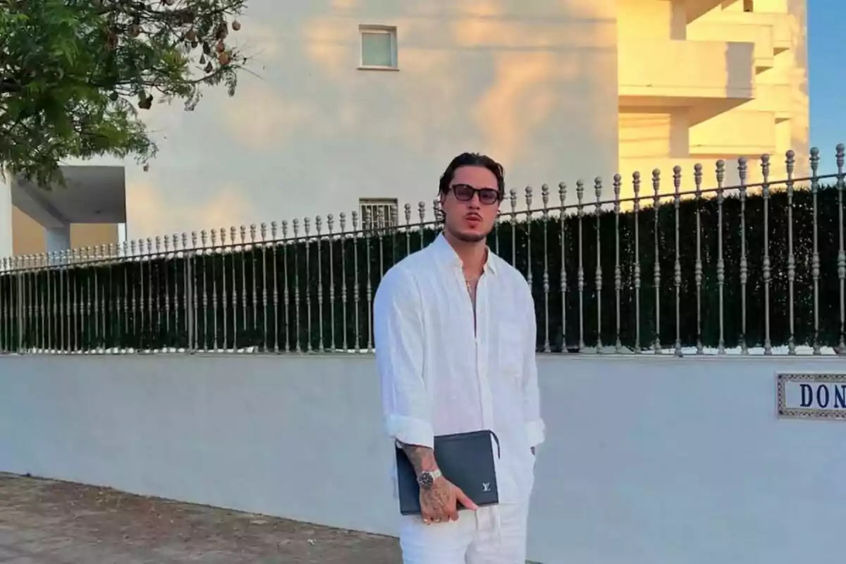 Carlo Costanzia vestido con camisa y pantalón blanco posando en la calle junto a una reja metálica y un edificio al fondo, sosteniendo una carpeta negra y usando gafas de sol. Carlo Costanzia vestido con camisa y pantalón blanco posando en la calle junto a una reja metálica y un edificio al fondo, sosteniendo una carpeta negra y usando gafas de sol.