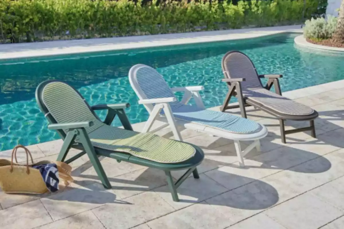 Tres tumbonas de diferentes colores están alineadas junto a una piscina con agua cristalina y al fondo se observa vegetación, mientras una bolsa de playa y un sombrero están en el suelo. Tres tumbonas de diferentes colores están alineadas junto a una piscina con agua cristalina y al fondo se observa vegetación, mientras una bolsa de playa y un sombrero están en el suelo.