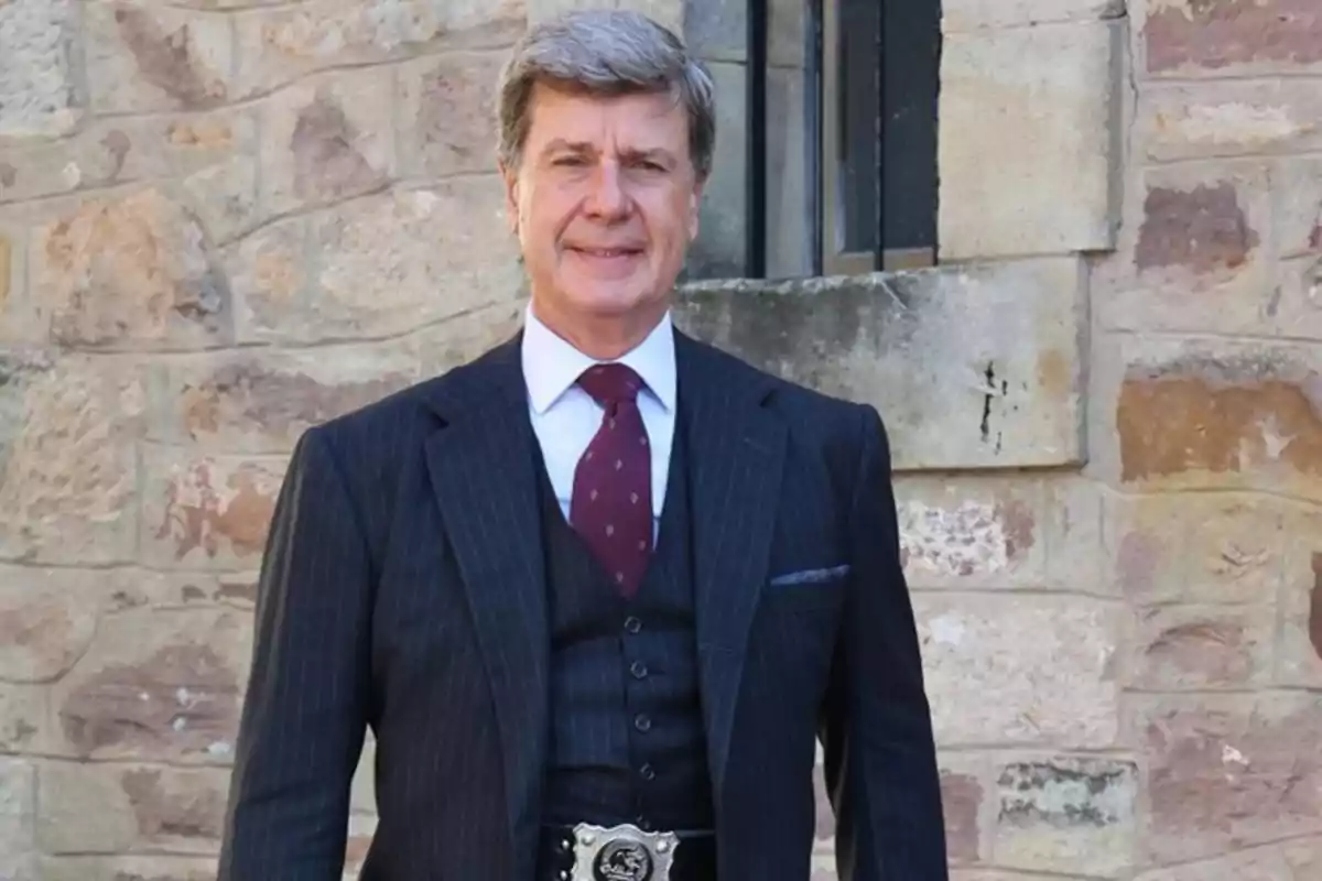 Hombre de cabello canoso vistiendo traje oscuro con corbata roja posando frente a una pared de piedra
