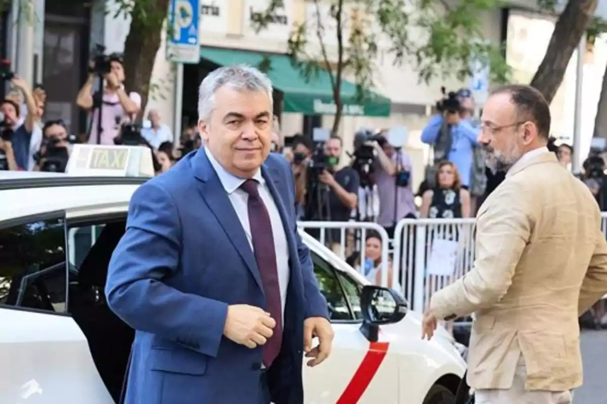 Un hombre con traje azul sale de un taxi blanco mientras varias personas y fotógrafos lo observan en la calle