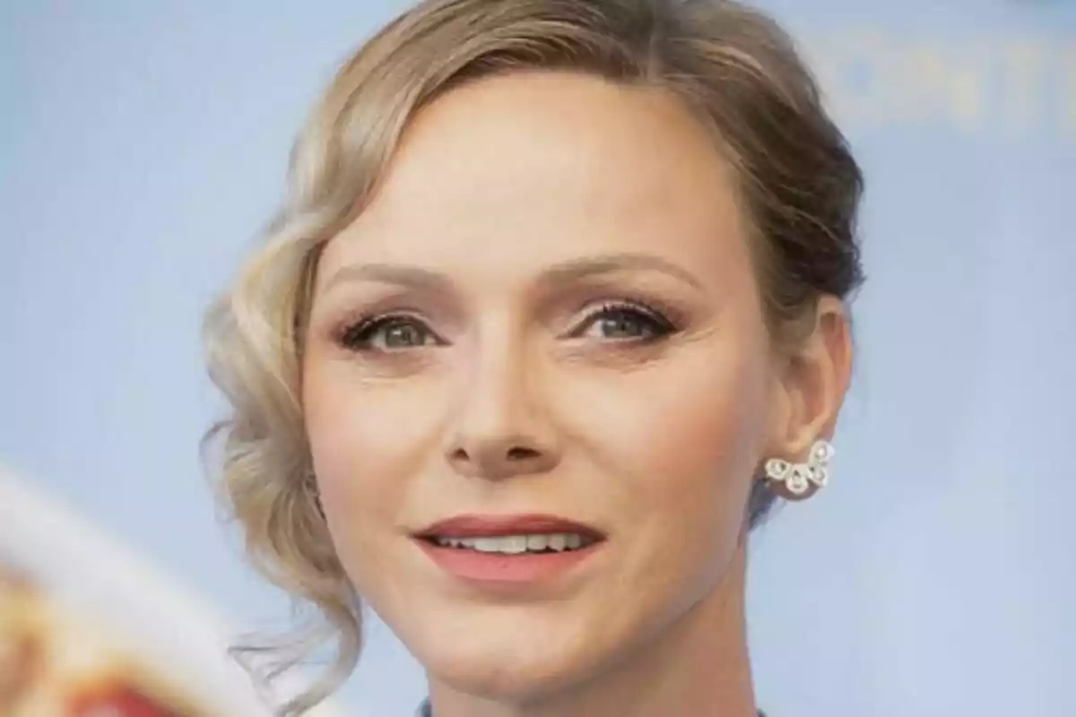 Primer plano de Charlene de Mónaco con maquillaje suave y aretes brillantes, fondo azul desenfocado.