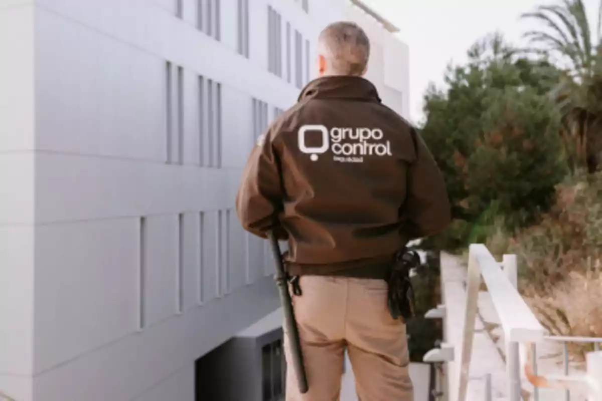 Guardia de seguridad de espaldas con chaqueta de Grupo Control en un entorno exterior junto a un edificio moderno y vegetación Guardia de seguridad de espaldas con chaqueta de Grupo Control en un entorno exterior junto a un edificio moderno y vegetación