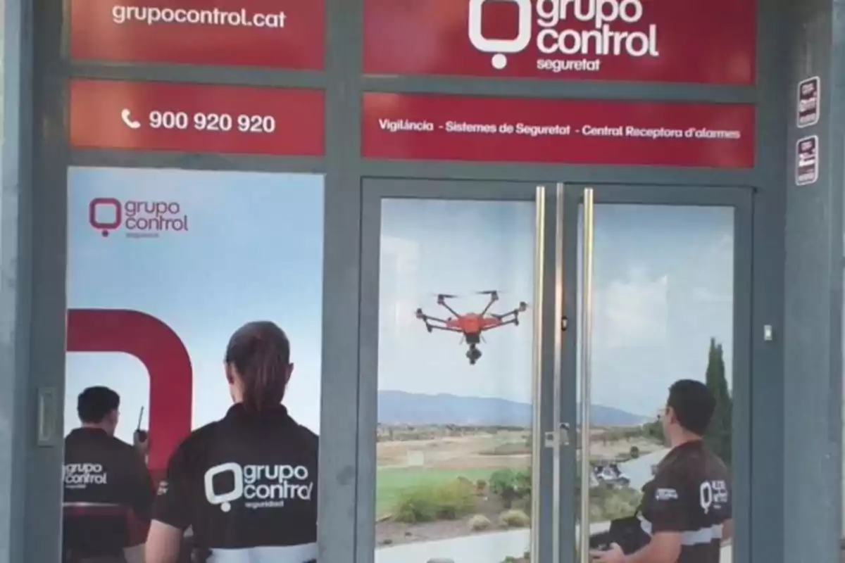 Entrada de una empresa de seguridad con imágenes de empleados y un dron en la puerta de cristal, junto a logotipos y datos de contacto de Grupo Control Entrada de una empresa de seguridad con imágenes de empleados y un dron en la puerta de cristal, junto a logotipos y datos de contacto de Grupo Control
