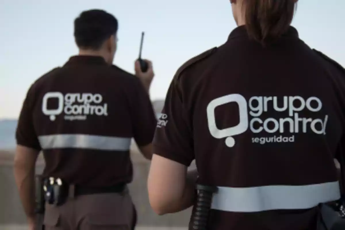 Dos personas con uniforme marrón de seguridad de Grupo Control están de espaldas, una sostiene un walkie talkie y ambas llevan cinturón con equipo.