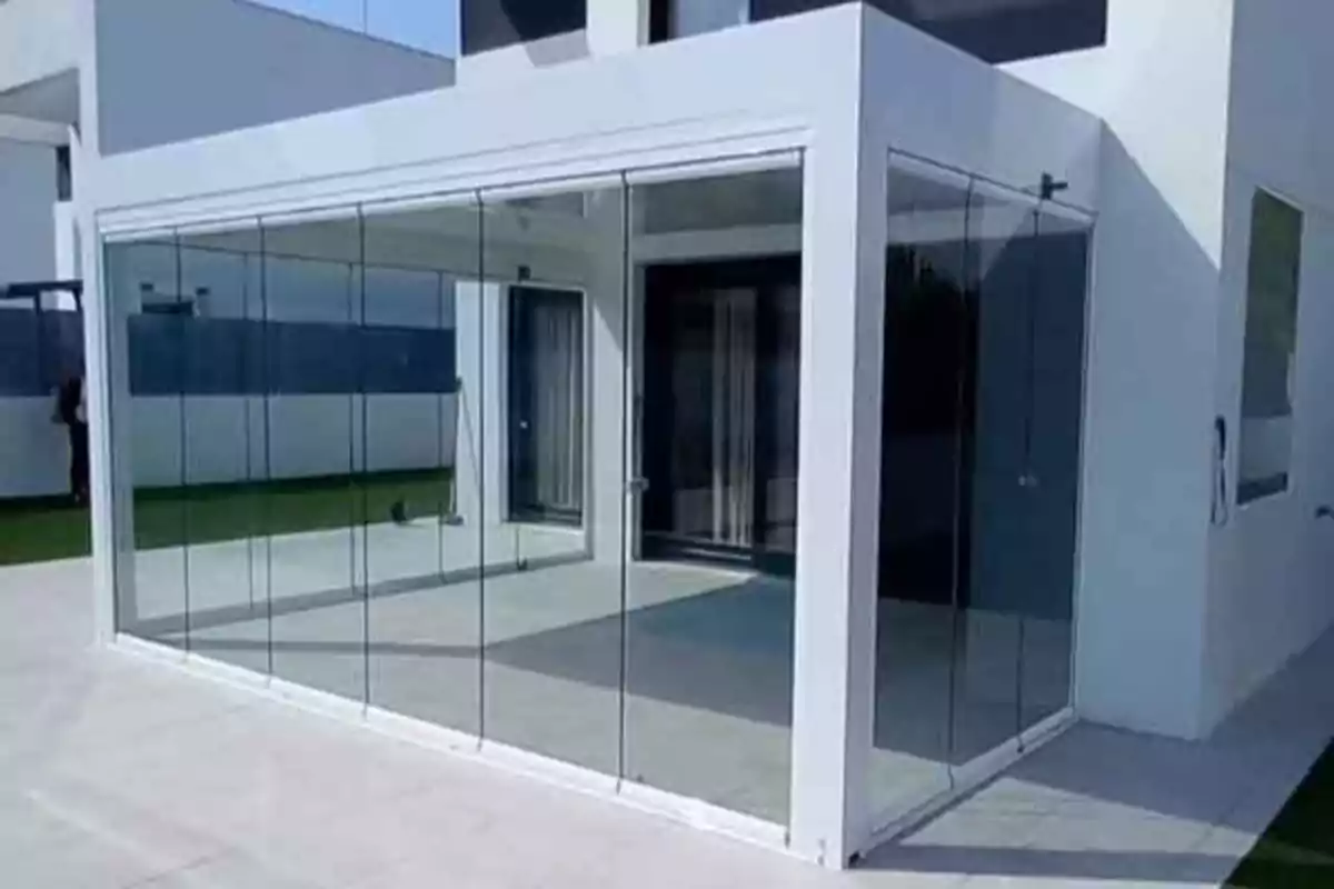 Estructura moderna de terraza cerrada con paneles de vidrio junto a una casa blanca