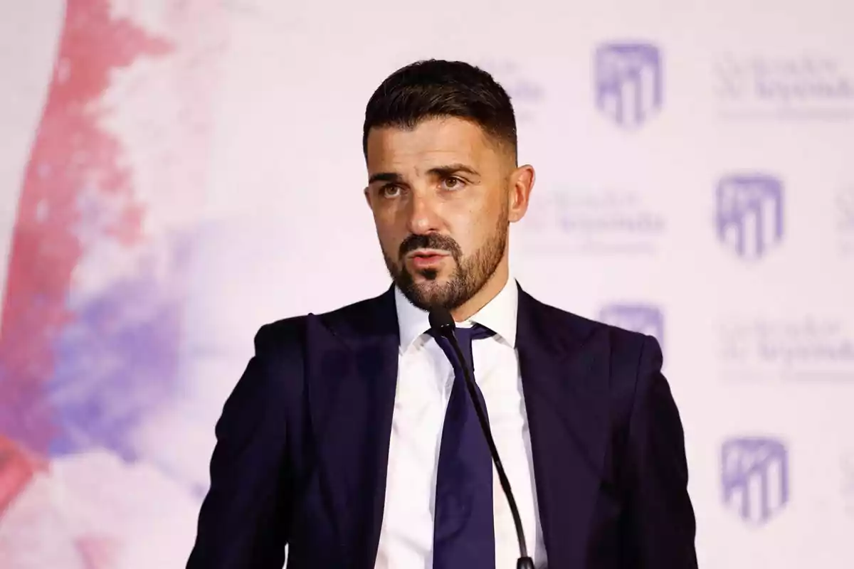 Hombre con barba y cabello corto vestido con traje oscuro y corbata hablando frente a un micrófono en un evento formal Hombre con barba y cabello corto vestido con traje oscuro y corbata hablando frente a un micrófono en un evento formal