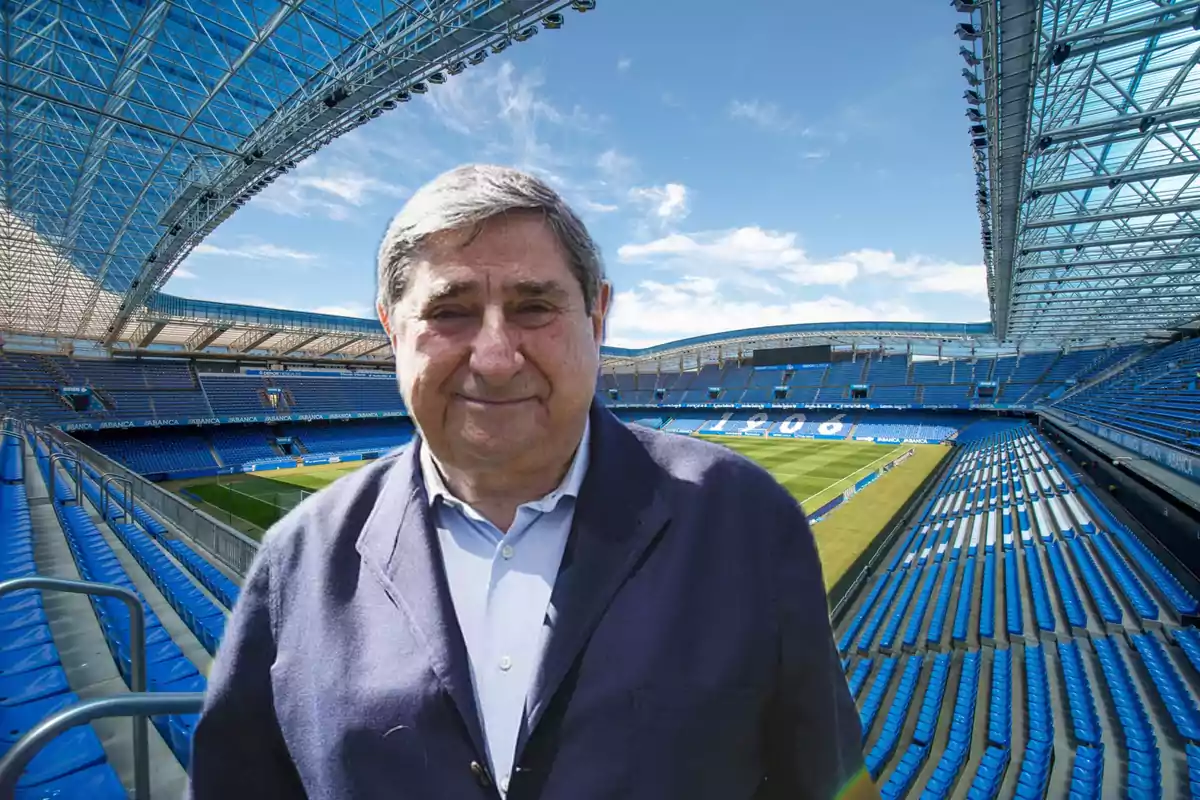 Hombre mayor de cabello canoso con chaqueta azul posando en un estadio de fútbol vacío con gradas azules y techo parcialmente cubierto