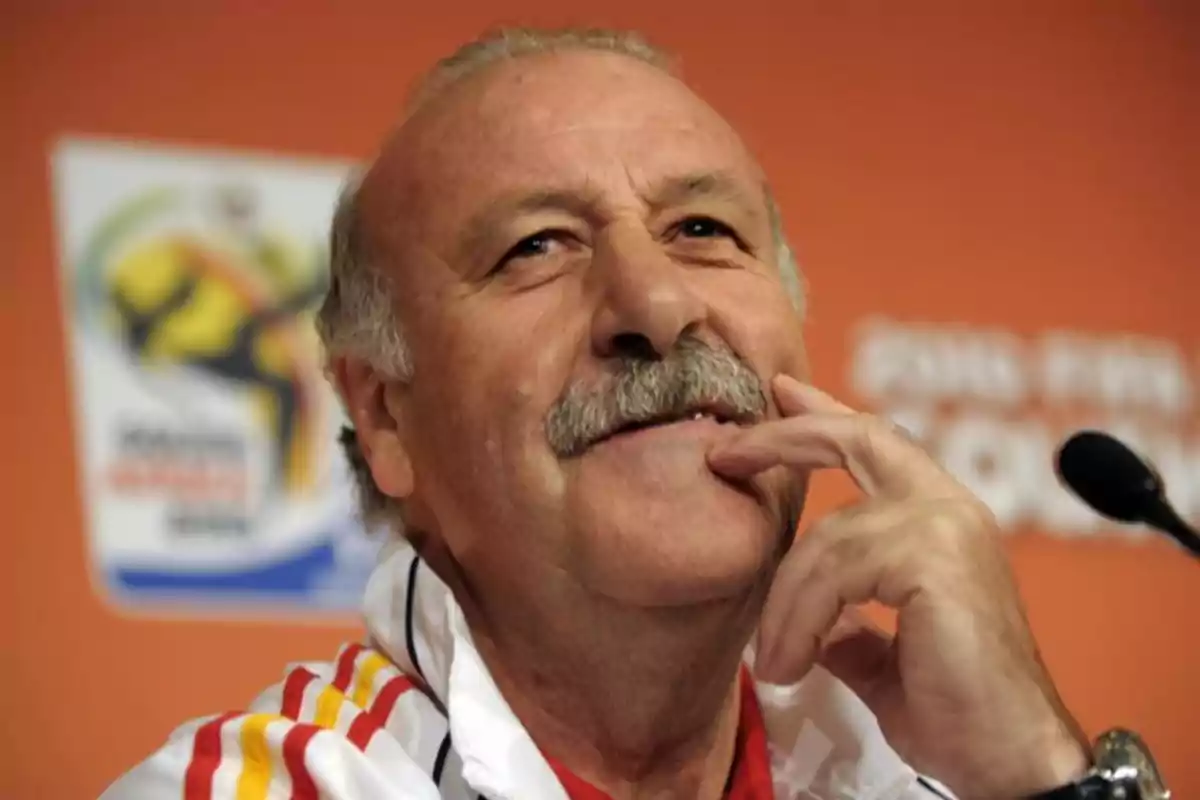 Hombre mayor con bigote sonríe pensativo durante una conferencia de prensa con fondo naranja y un micrófono a un lado Hombre mayor con bigote sonríe pensativo durante una conferencia de prensa con fondo naranja y un micrófono a un lado