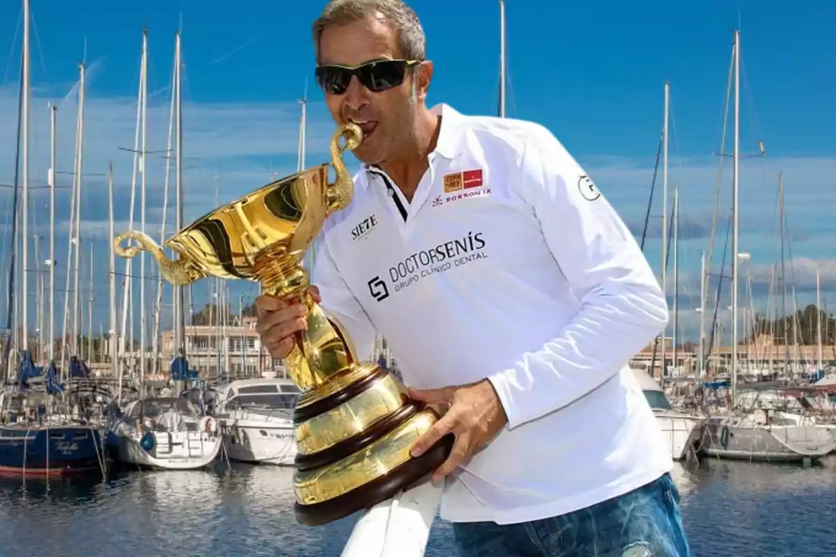 Hombre con gafas de sol y camiseta blanca celebrando con un trofeo dorado en un puerto deportivo lleno de veleros Hombre con gafas de sol y camiseta blanca celebrando con un trofeo dorado en un puerto deportivo lleno de veleros