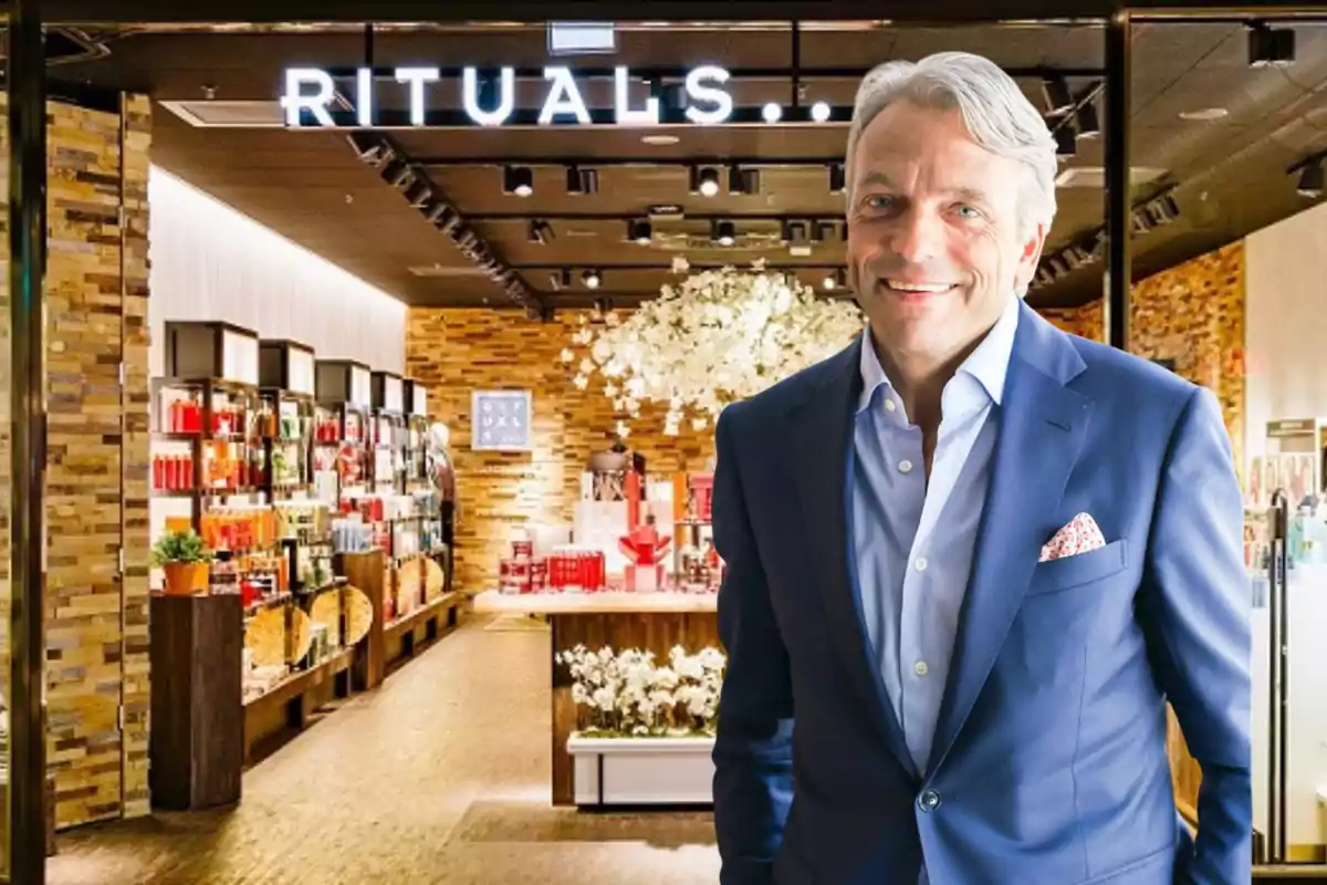 Hombre de cabello canoso con traje azul sonriendo frente a la entrada de una tienda Rituals iluminada y decorada con productos y flores blancas
