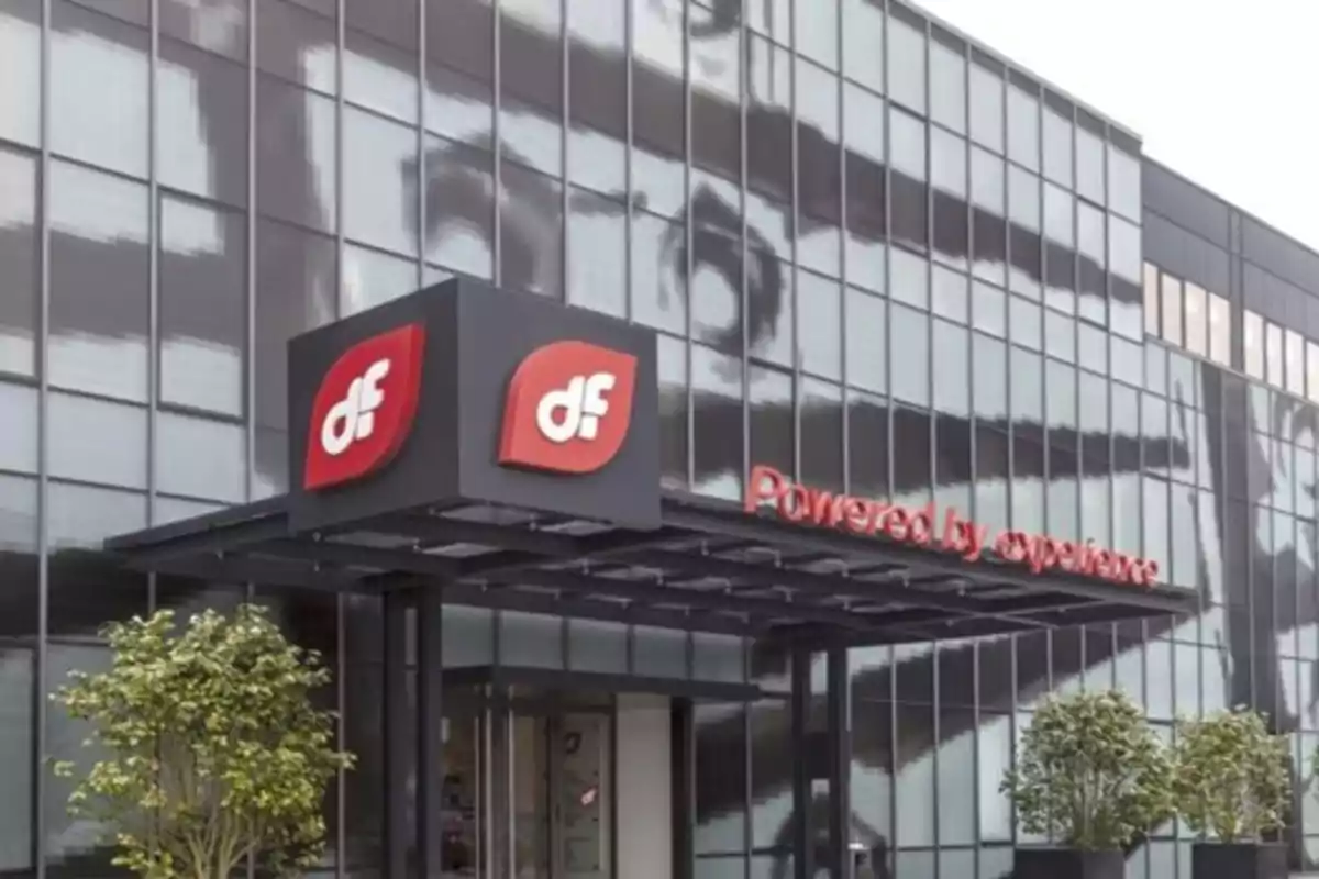 Fachada de edificio moderno con logotipo rojo de df y letrero que dice Powered by experience junto a la entrada principal rodeada de plantas Fachada de edificio moderno con logotipo rojo de df y letrero que dice Powered by experience junto a la entrada principal rodeada de plantas