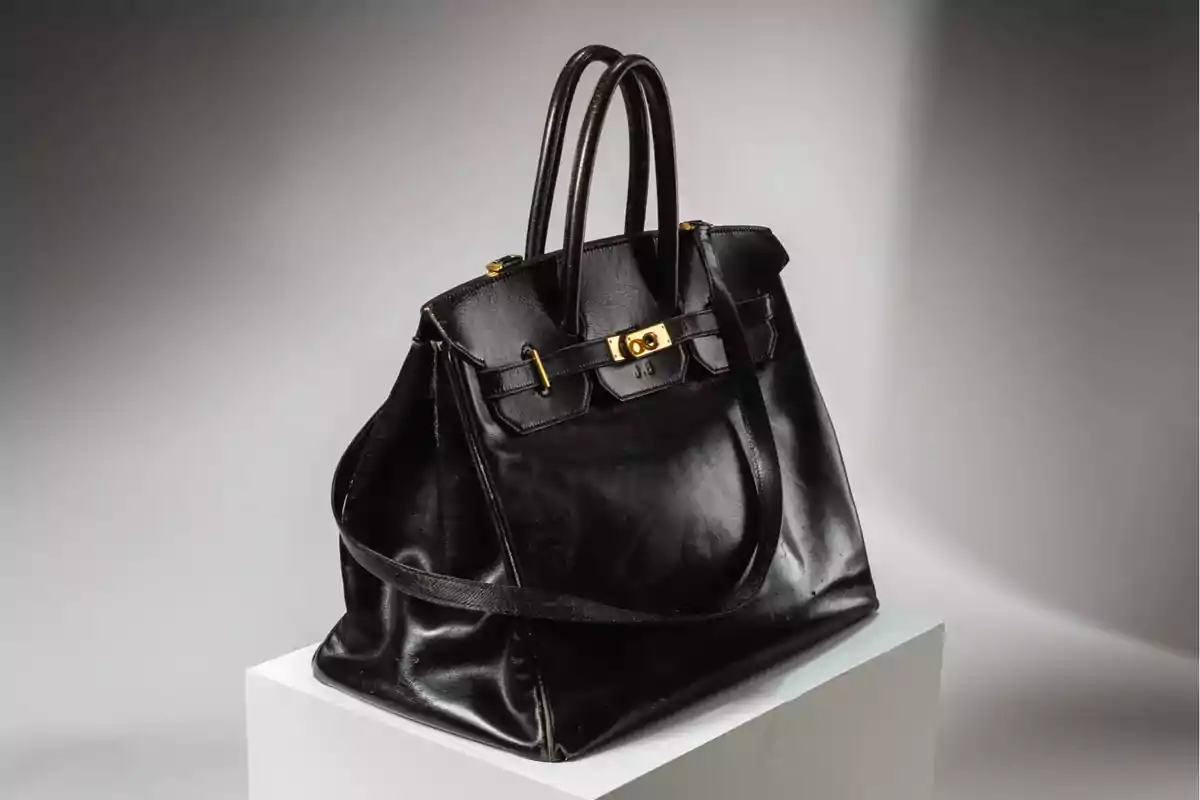 Bolso de cuero negro con herrajes dorados y doble asa sobre un pedestal blanco
