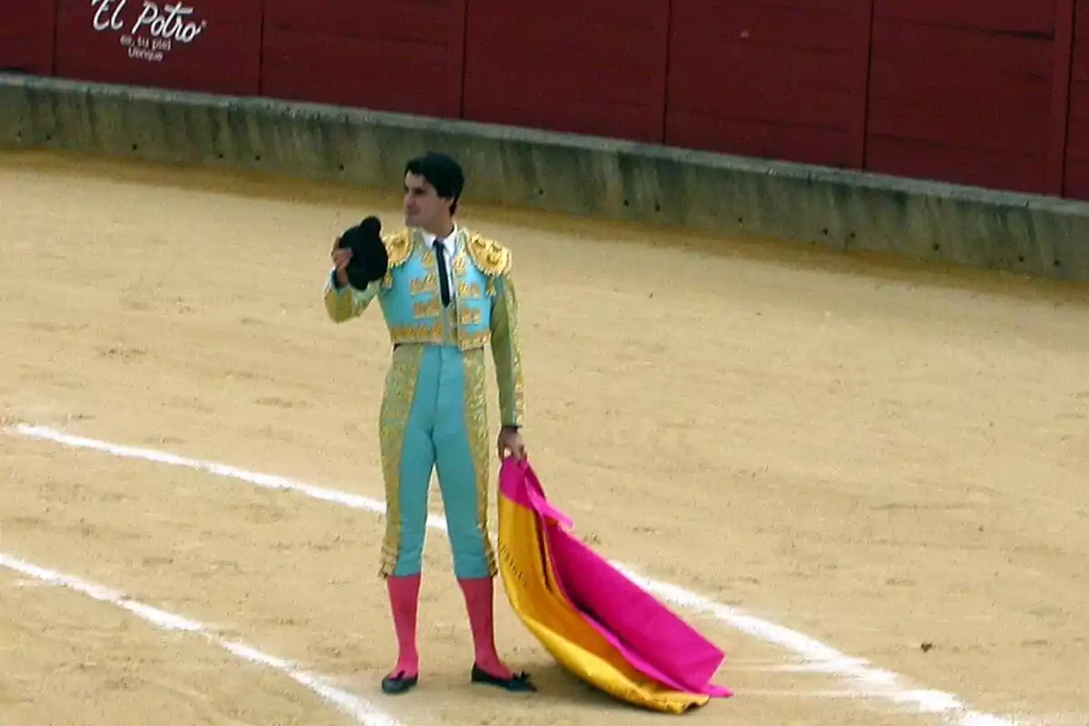 Un torero vestido con traje tradicional sostiene una montera en una mano y una capa en la otra dentro de una plaza de toros Un torero vestido con traje tradicional sostiene una montera en una mano y una capa en la otra dentro de una plaza de toros