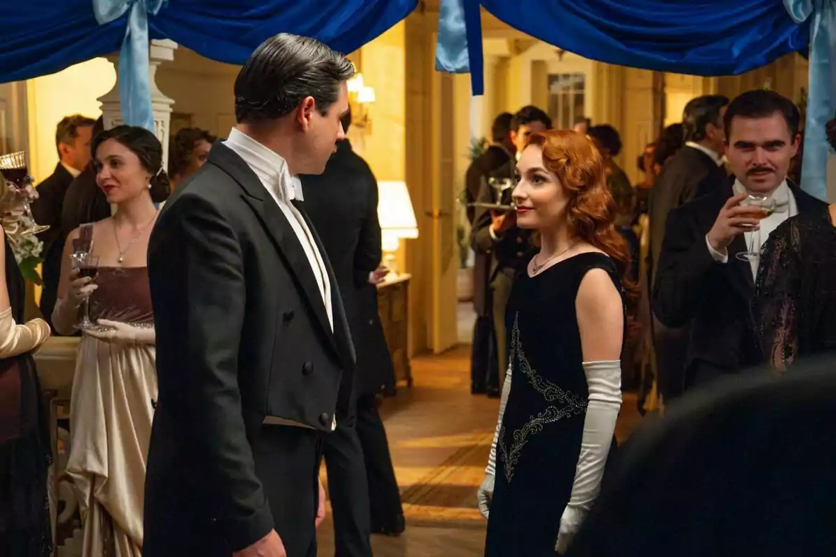 Dos personas -que son Arturo Sancho y Sara Font- elegantemente vestidas conversan en una fiesta de época decorada con cortinas azules. Dos personas -que son Arturo Sancho y Sara Font- elegantemente vestidas conversan en una fiesta de época decorada con cortinas azules.