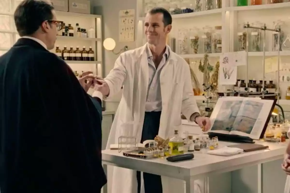 Un hombre con bata blanca sonríe mientras entrega un frasco pequeño a otra persona en un laboratorio lleno de frascos y plantas secas, se trata de una escena de la serie 'Sueños de Libertad'. Un hombre con bata blanca sonríe mientras entrega un frasco pequeño a otra persona en un laboratorio lleno de frascos y plantas secas, se trata de una escena de la serie 'Sueños de Libertad'.