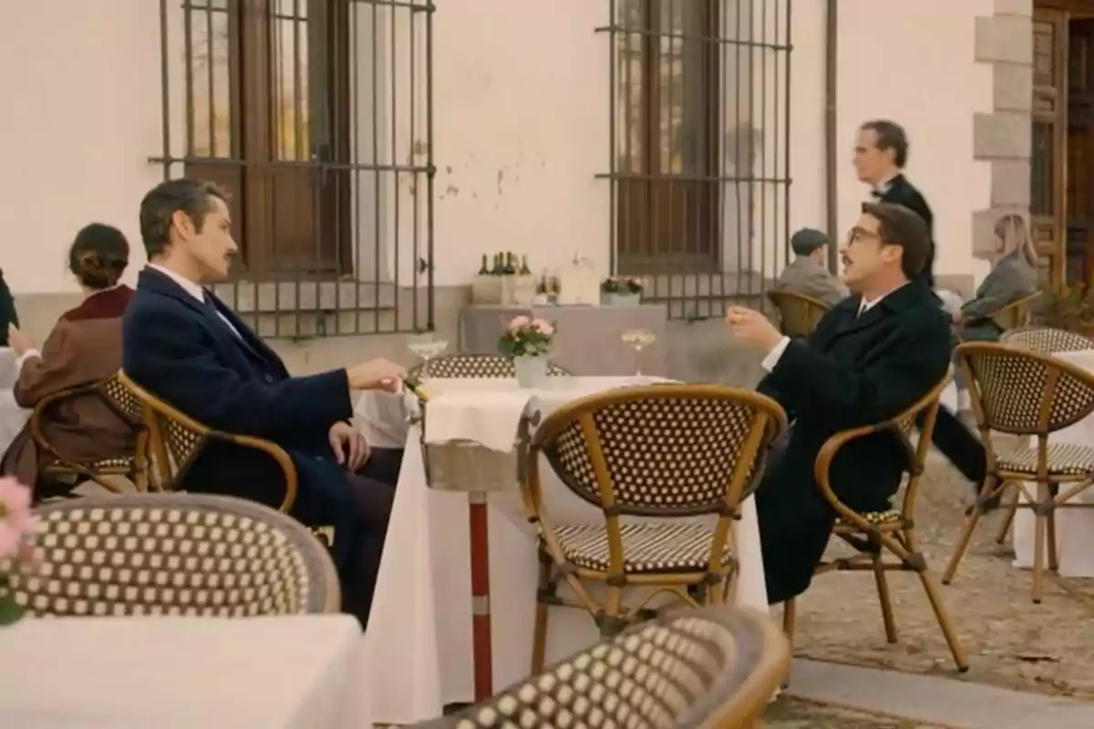 Dos hombres con bigote conversan sentados en una terraza de cafetería al aire libre en una escena de 'Sueños de Libertad', rodeados de mesas con manteles blancos y flores, mientras otras personas están en el fondo. Dos hombres con bigote conversan sentados en una terraza de cafetería al aire libre en una escena de 'Sueños de Libertad', rodeados de mesas con manteles blancos y flores, mientras otras personas están en el fondo.