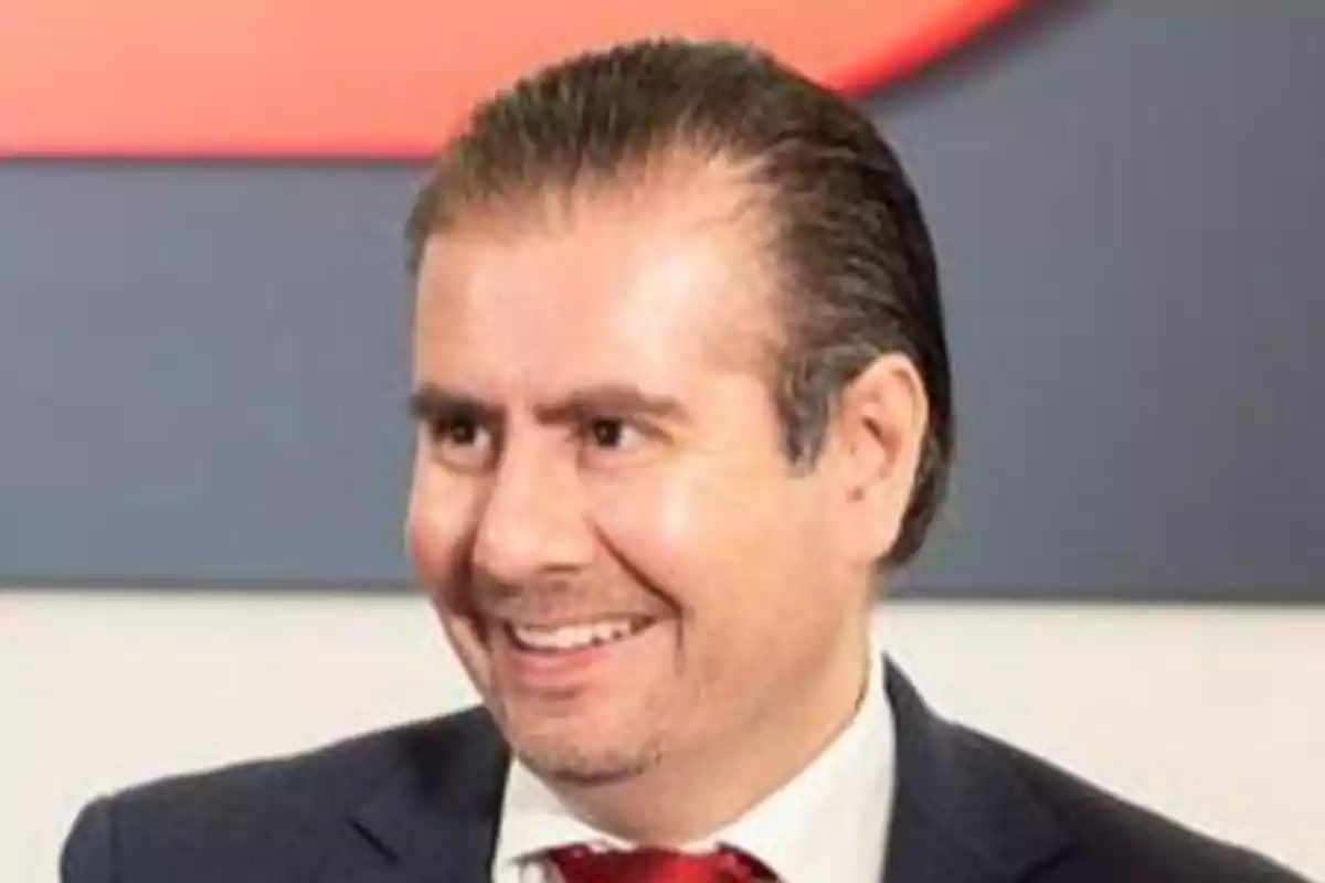 Hombre de cabello oscuro y peinado hacia atrás, vestido con traje y corbata roja, sonriendo en un entorno interior. Hombre de cabello oscuro y peinado hacia atrás, vestido con traje y corbata roja, sonriendo en un entorno interior.