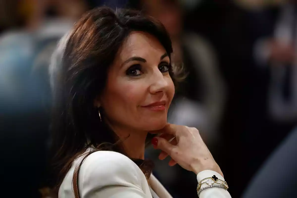Mujer de cabello castaño oscuro y ojos claros con expresión sonriente, vestida con saco blanco y accesorios dorados, mirando hacia la cámara en un ambiente interior desenfocado.