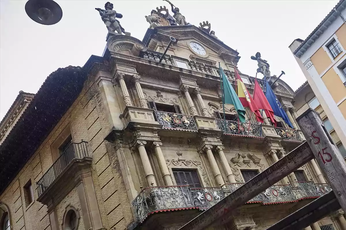 Fachada de un edificio histórico con balcones de hierro forjado, columnas ornamentadas, varias banderas y estatuas en la parte superior Fachada de un edificio histórico con balcones de hierro forjado, columnas ornamentadas, varias banderas y estatuas en la parte superior