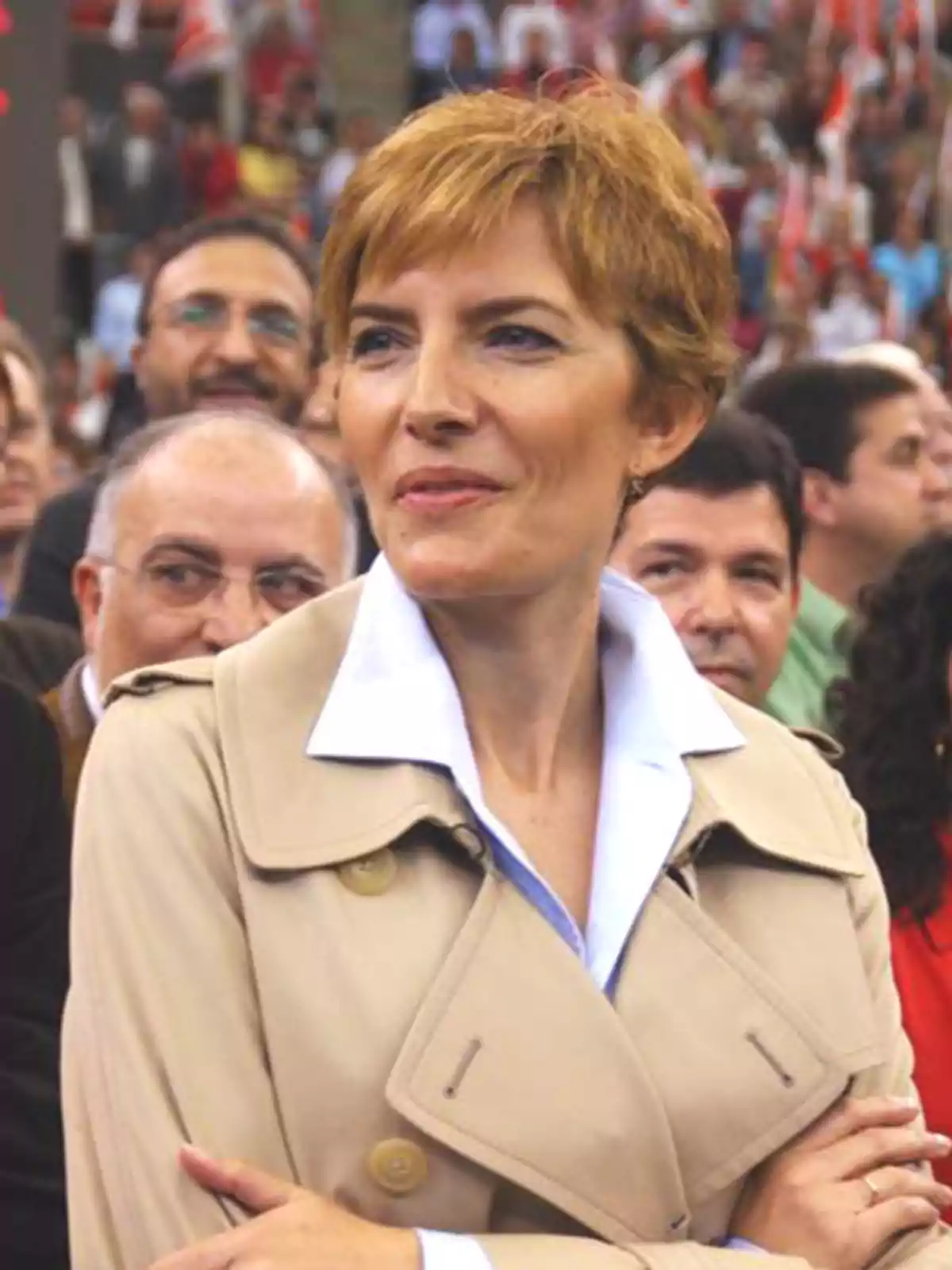 Mujer de cabello corto y rubio con gabardina beige y camisa blanca rodeada de personas en un evento al aire libre