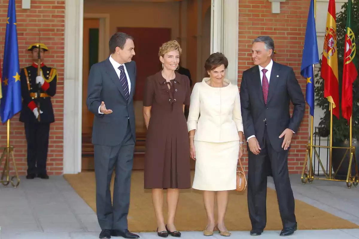 Cuatro personas elegantemente vestidas posan sonrientes en la entrada de un edificio oficial con banderas de la Unión Europea, España y Portugal al fondo y un guardia uniformado a la izquierda