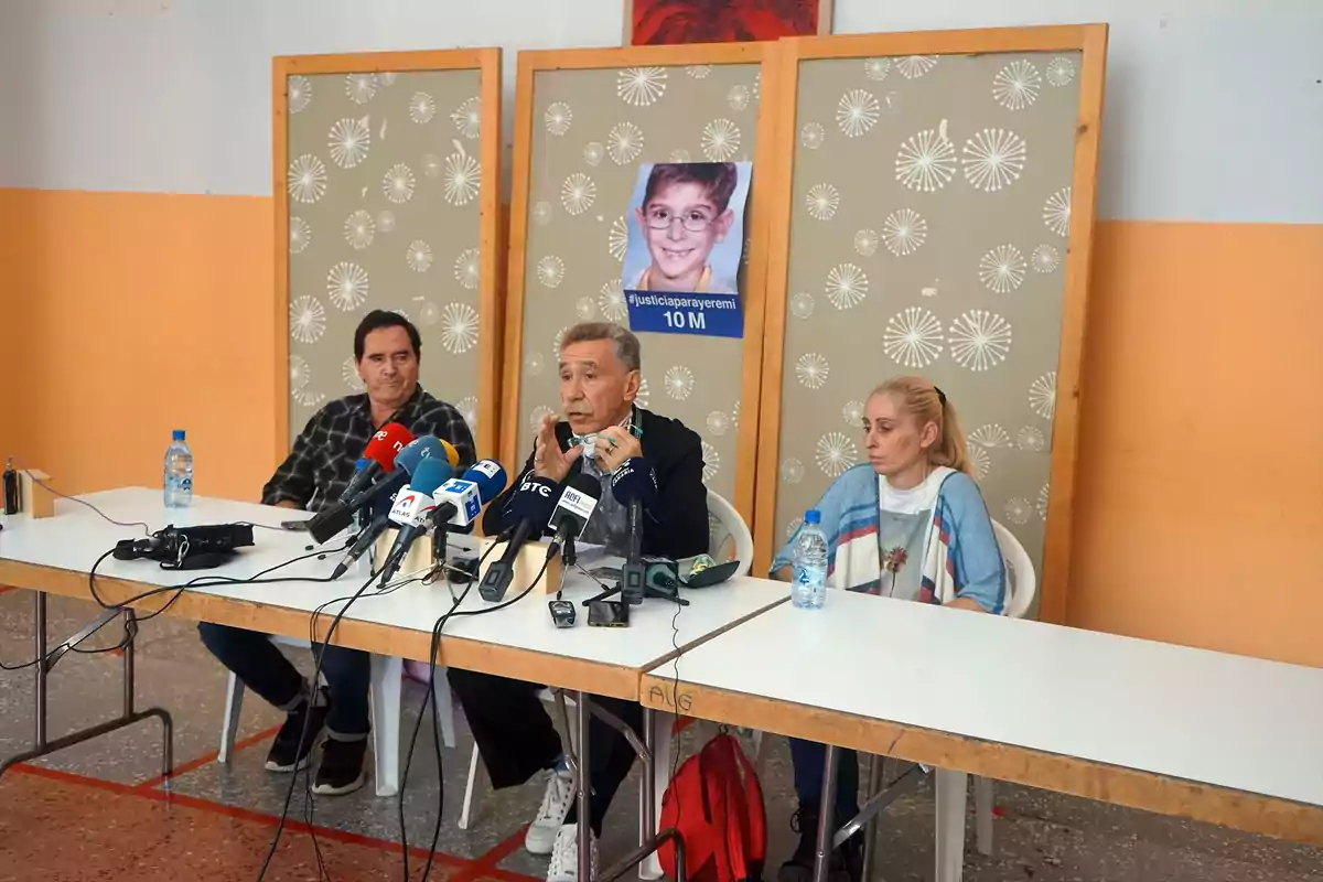 Tres personas sentadas en una mesa durante una rueda de prensa con varios micrófonos frente a ellas y una foto de un niño con gafas pegada en un panel detrás