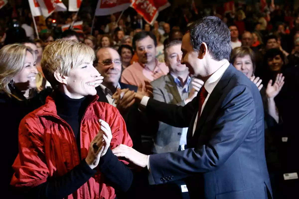 Un hombre con traje saluda a una mujer sonriente vestida con chaqueta roja mientras un grupo de personas aplaude y sostiene banderas en el fondo