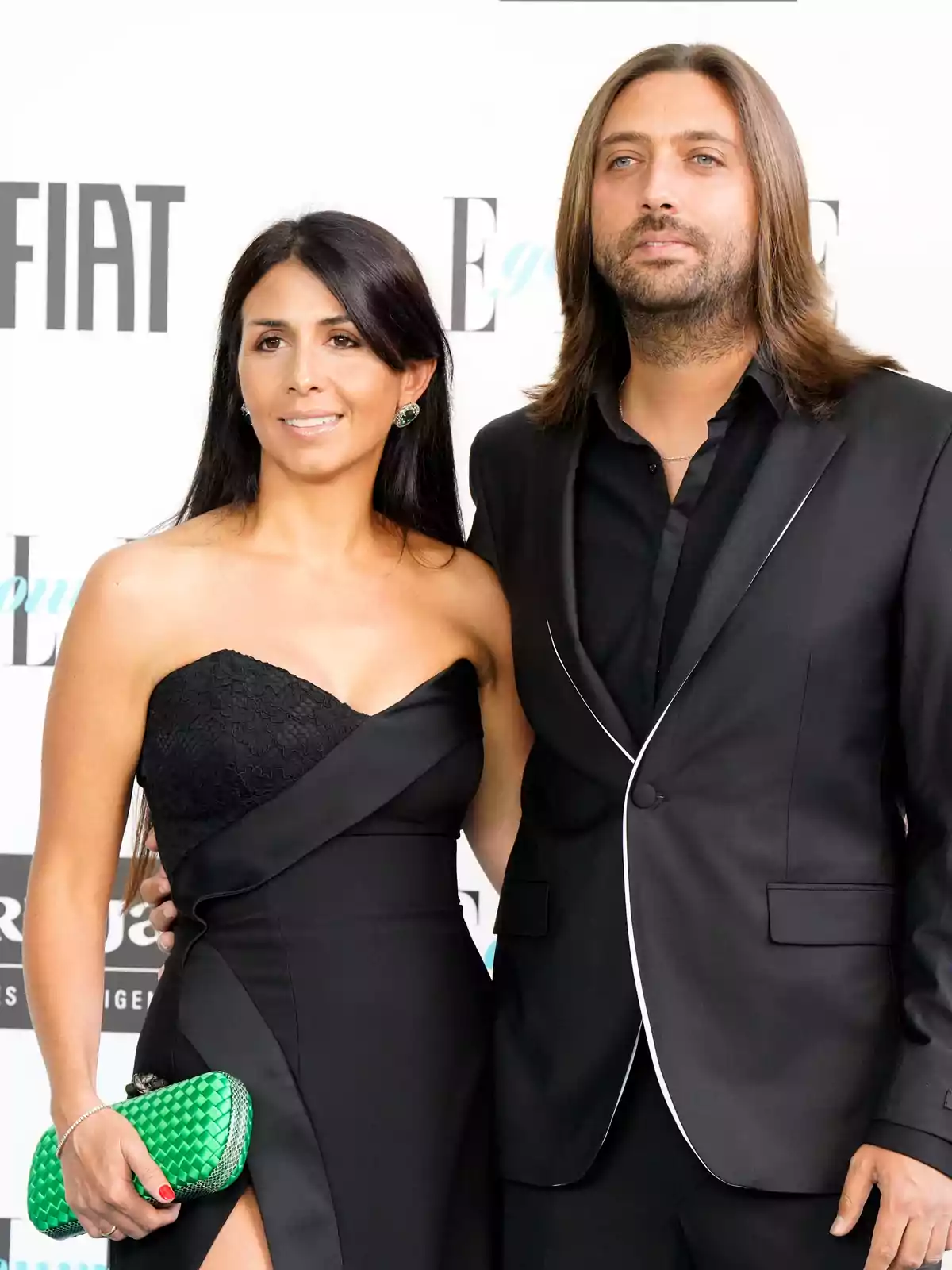 Pareja posando en un evento elegante ambos vestidos de negro ella lleva un bolso verde y un vestido con abertura en la pierna