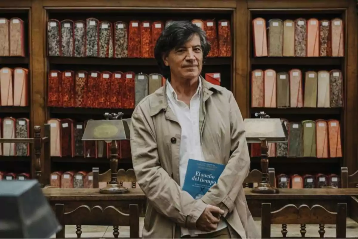 Hombre de cabello canoso y abrigo beige sostiene un libro azul en una biblioteca antigua con estanterías llenas de archivos y lámparas de escritorio de fondo Hombre de cabello canoso y abrigo beige sostiene un libro azul en una biblioteca antigua con estanterías llenas de archivos y lámparas de escritorio de fondo