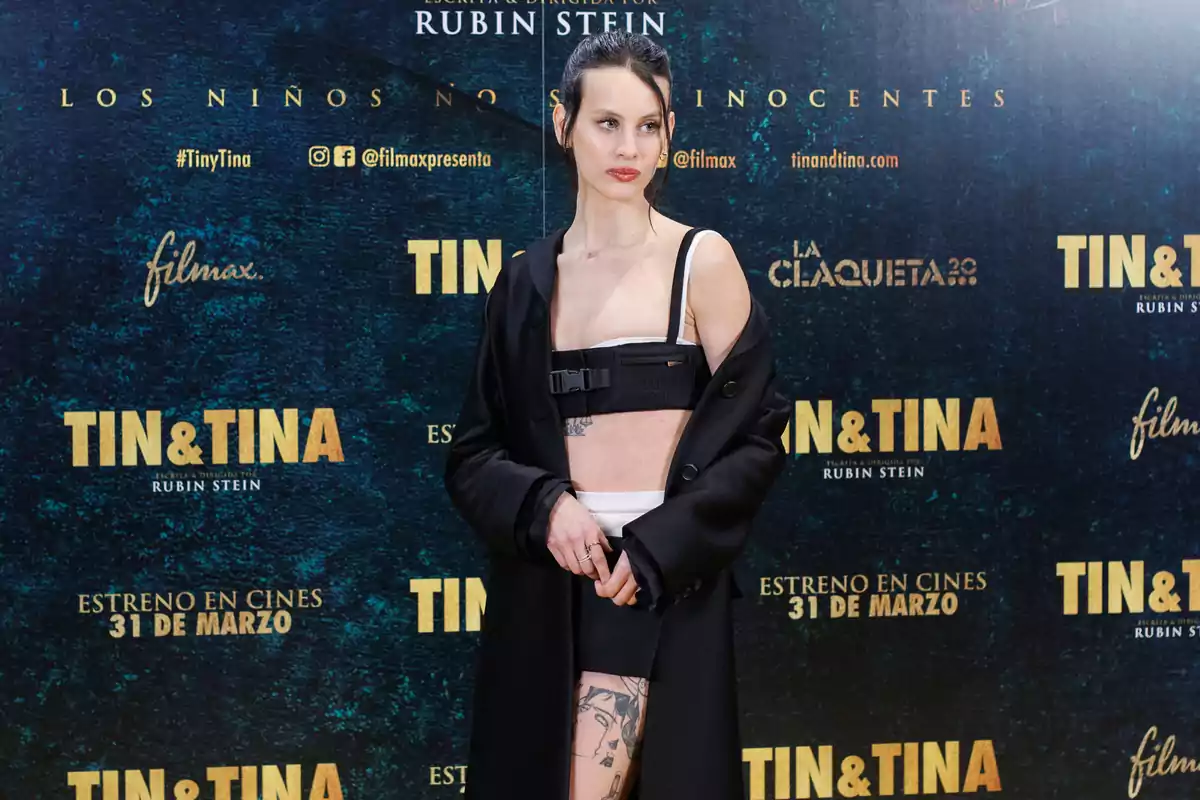 Mujer posando en la alfombra roja de un evento de estreno de la película Tin & Tina, vistiendo un abrigo negro y un conjunto moderno con tatuajes visibles en la pierna y el torso, sobre un fondo promocional azul oscuro con letras doradas. Mujer posando en la alfombra roja de un evento de estreno de la película Tin & Tina, vistiendo un abrigo negro y un conjunto moderno con tatuajes visibles en la pierna y el torso, sobre un fondo promocional azul oscuro con letras doradas.