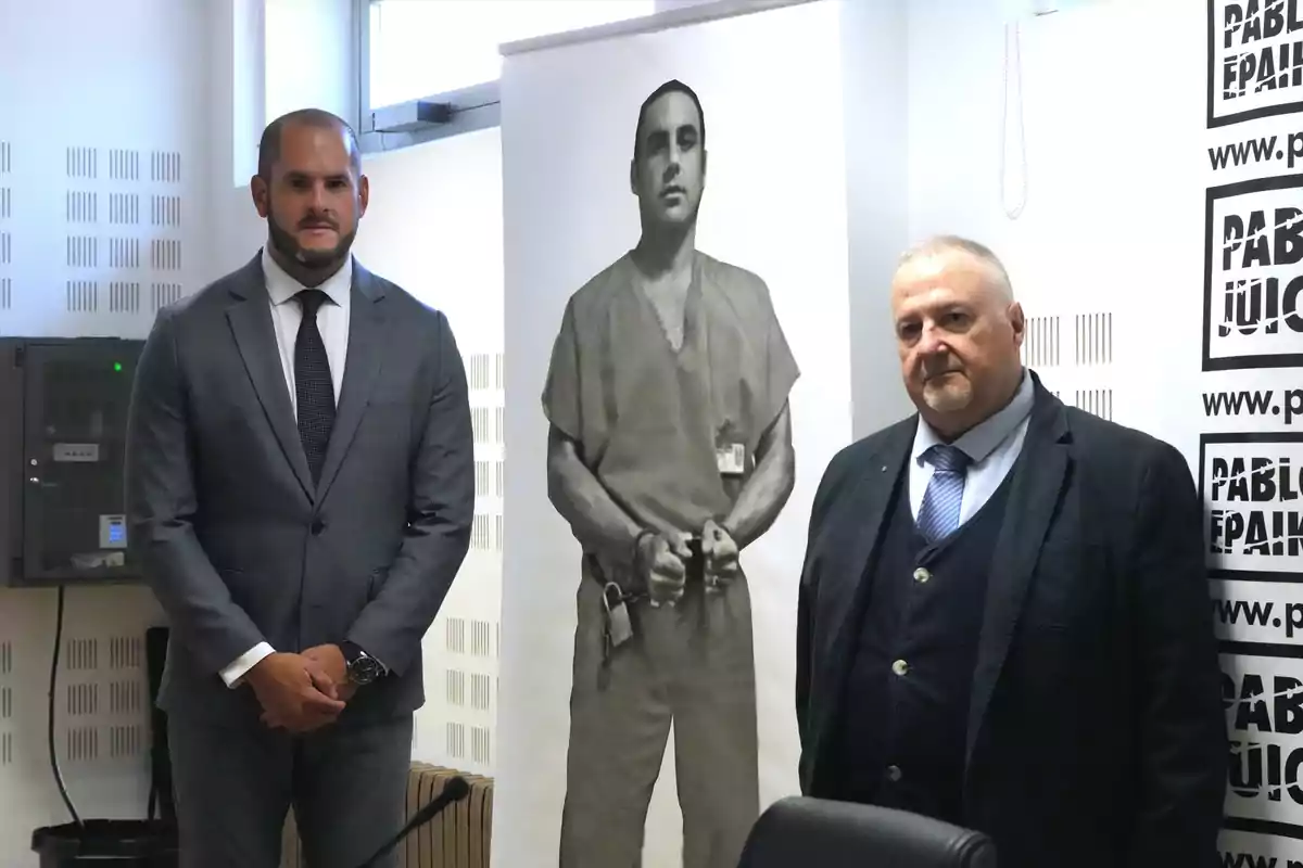 Dos hombres de traje posan junto a una imagen en blanco y negro de una persona esposada en un cartel durante un evento en interiores Dos hombres de traje posan junto a una imagen en blanco y negro de una persona esposada en un cartel durante un evento en interiores