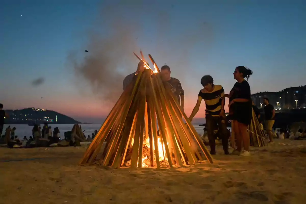 Personas encendiendo una hoguera en la playa al atardecer mientras otras personas disfrutan del ambiente festivo cerca del mar Personas encendiendo una hoguera en la playa al atardecer mientras otras personas disfrutan del ambiente festivo cerca del mar