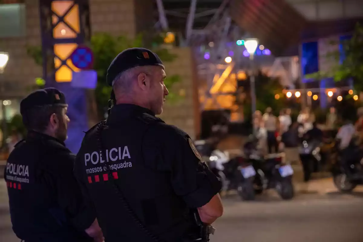 Dos agentes de la policía Mossos d'Esquadra vigilan una calle por la noche mientras varias personas se agrupan al fondo cerca de motocicletas y luces urbanas Dos agentes de la policía Mossos d'Esquadra vigilan una calle por la noche mientras varias personas se agrupan al fondo cerca de motocicletas y luces urbanas