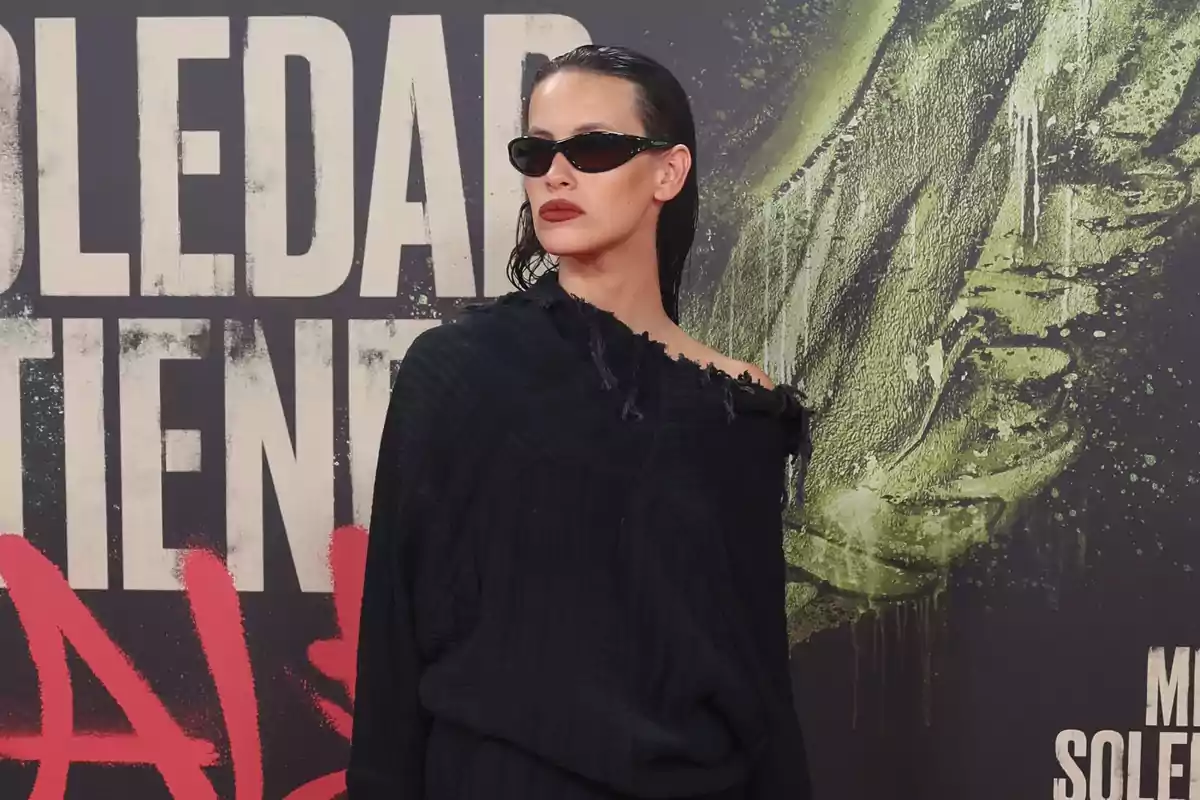 Mujer con gafas de sol y labios oscuros viste un suéter negro de hombro caído posando frente a un fondo con letras grandes y arte urbano. Mujer con gafas de sol y labios oscuros viste un suéter negro de hombro caído posando frente a un fondo con letras grandes y arte urbano.