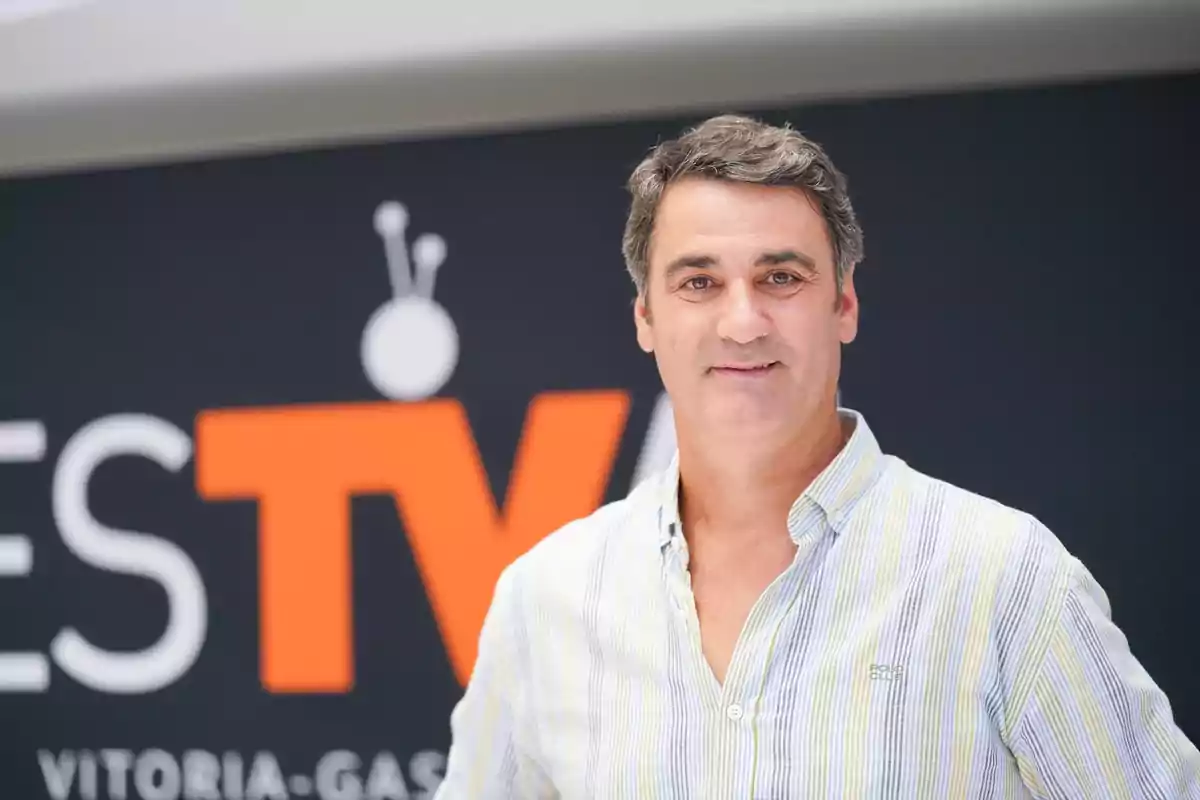 Hombre de cabello corto y canoso con camisa de rayas claras posando frente a un fondo con el logo de un canal de televisión