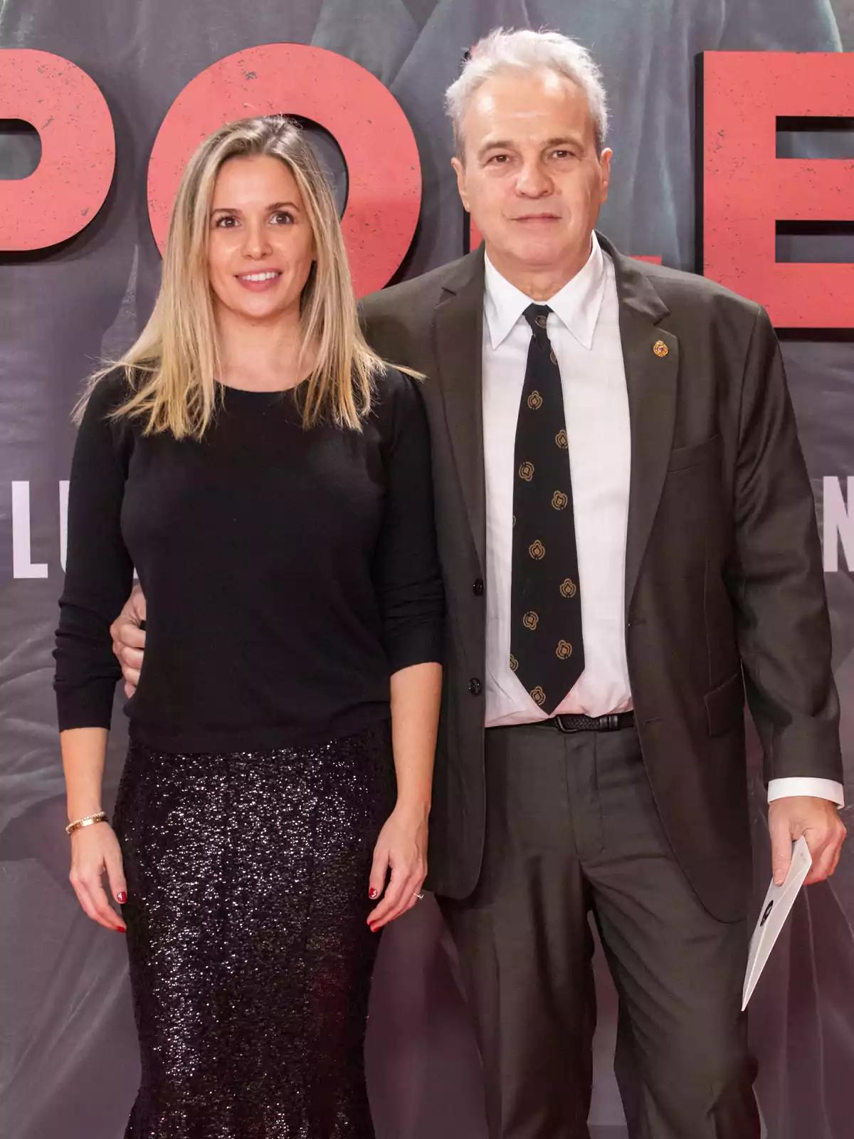 Una mujer con falda de lentejuelas negras y un hombre con traje posan juntos en un evento frente a un fondo con letras rojas grandes